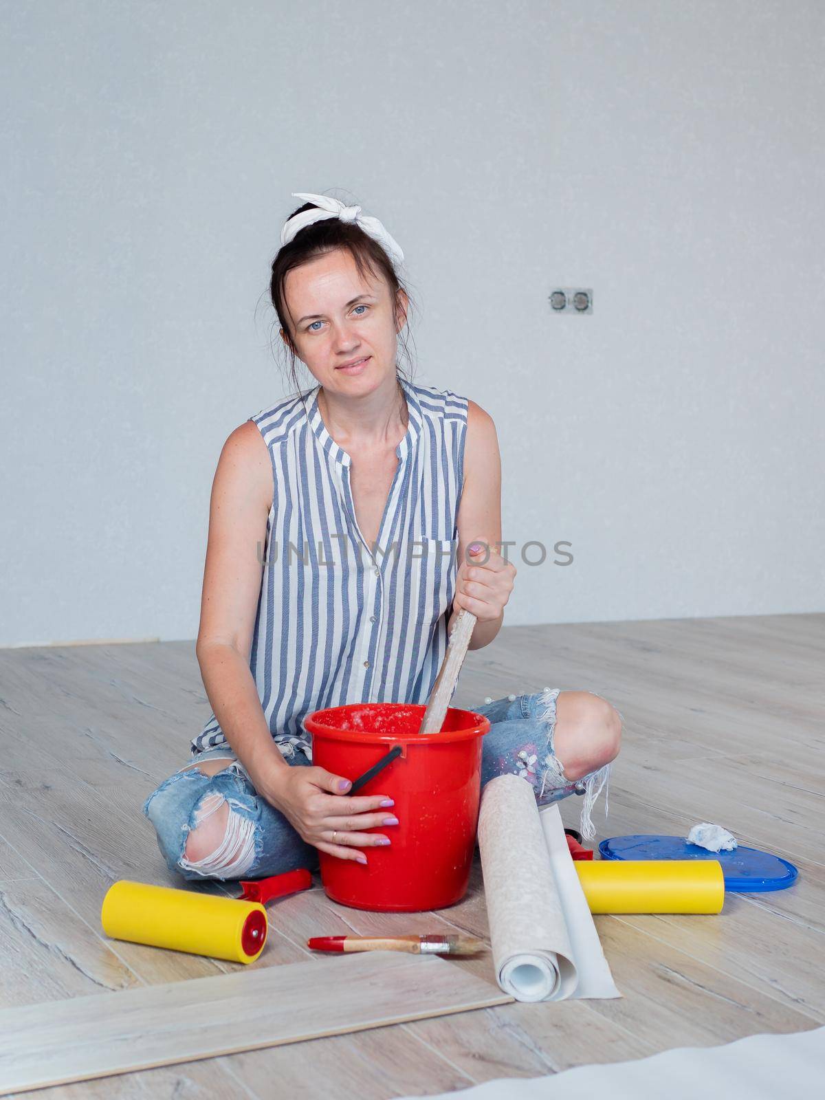 Portrait of a woman sitting on the floor of a room and stirring Wallpaper glue in a bucket. Concept of renovation in the apartment. by Utlanov