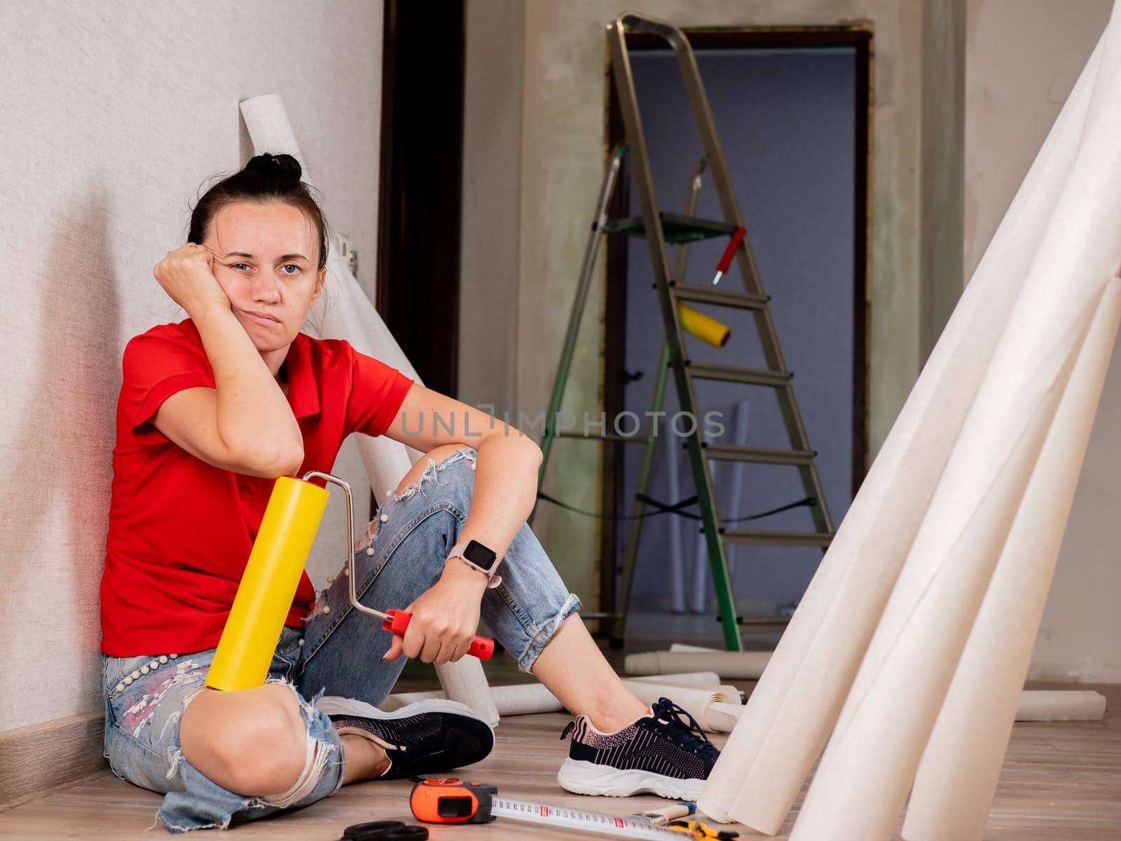 A woman sits on the floor and is sad because of the heavy repairs in the apartment. Wallpapering in the room. by Utlanov