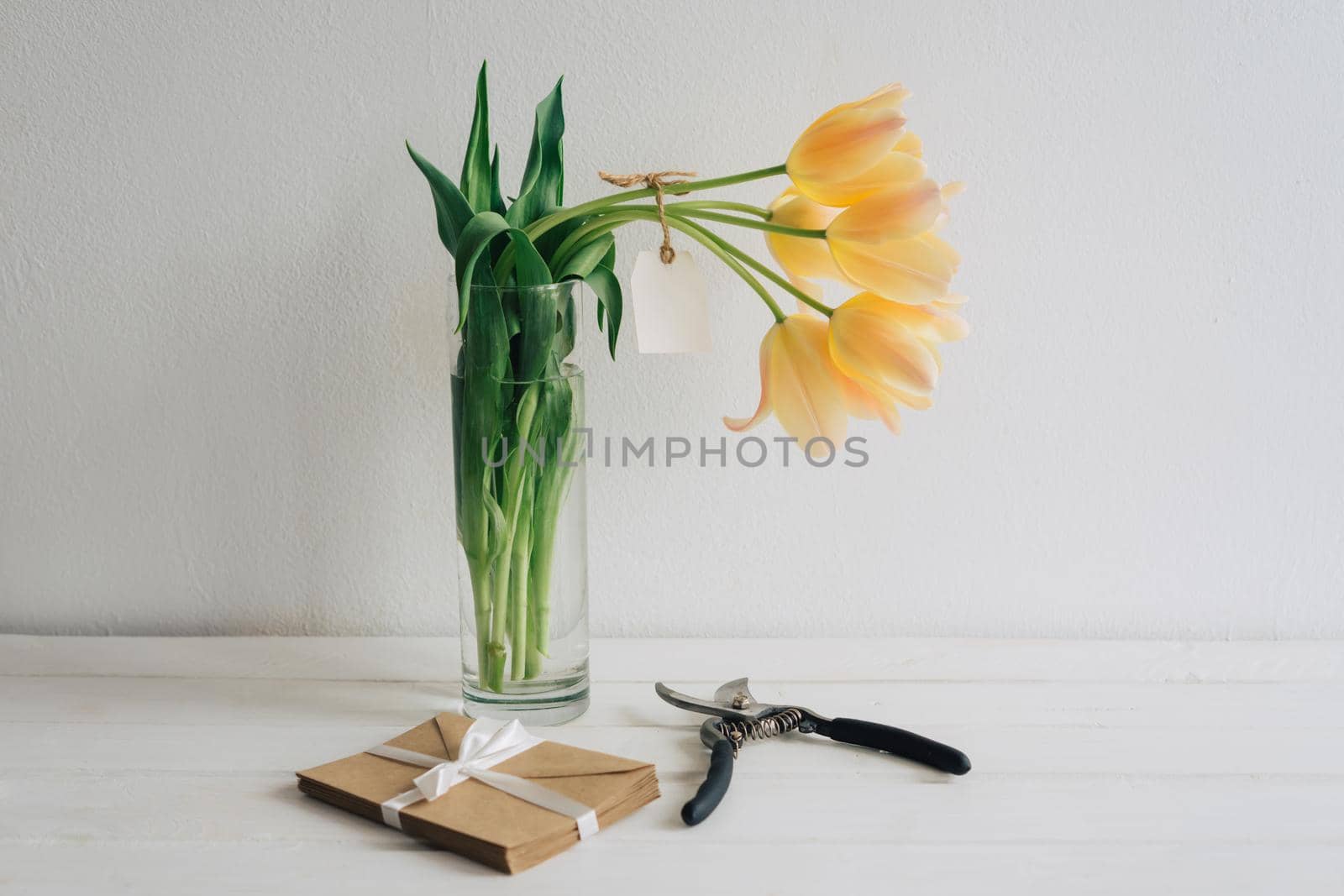 The secant-cut fragrant tulips stand in a glass vase. The tag is tied to a bouquet of yellow tulips. The envelopes of kraft paper are tied with a white satin ribbon. White wooden table.