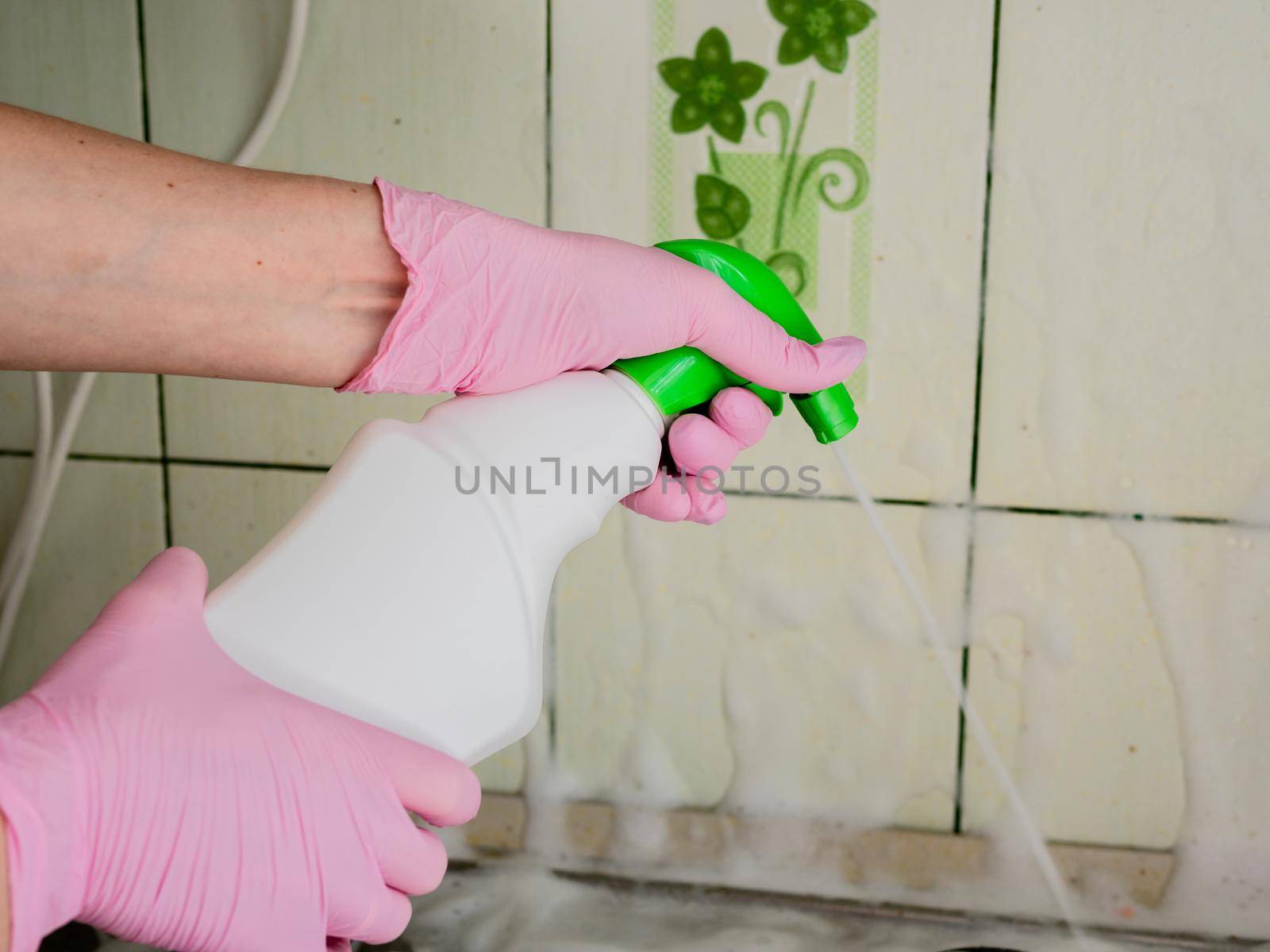 A woman in pink gloves sprays detergent on the tile above the gas stove to dissolve the fat and wash the dirty surface. A woman washes the tile above the kitchen stove of fat.