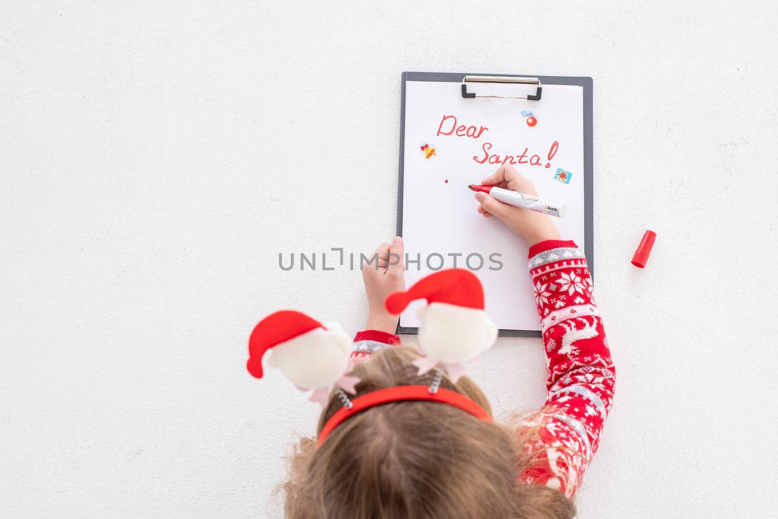 Dear Santa letter, Christmas card. Cute young girl wearing deer horns, holding a pen and writing on a white sheet