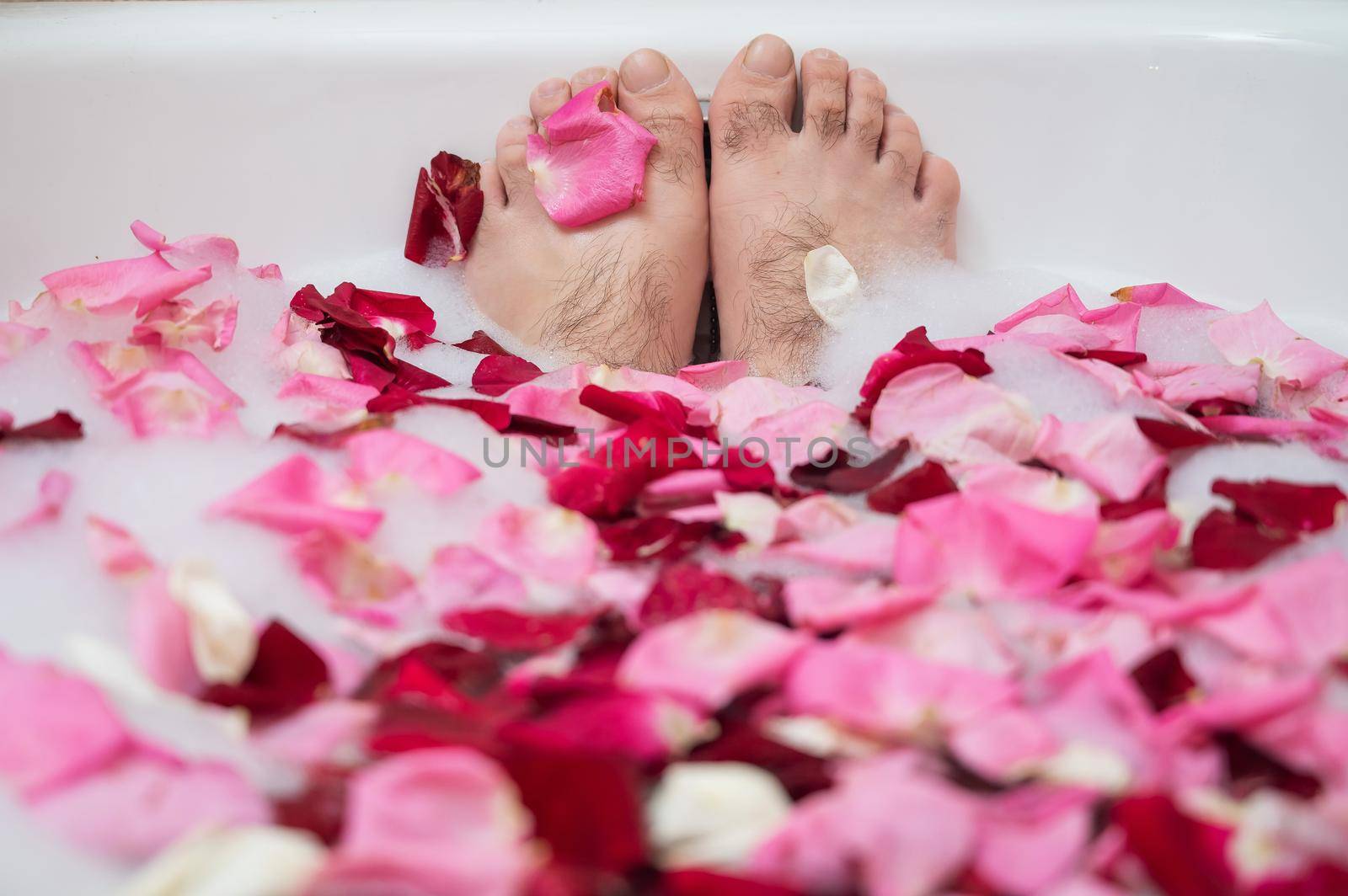 Funny picture of a man taking a relaxing bath. Close-up of male feet in a bath with foam and rose petals.