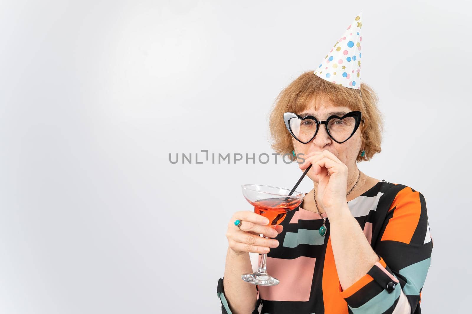 Portrait of a smiling elderly woman in a festive cap and glasses with hearts drinking a cocktail on a white background.