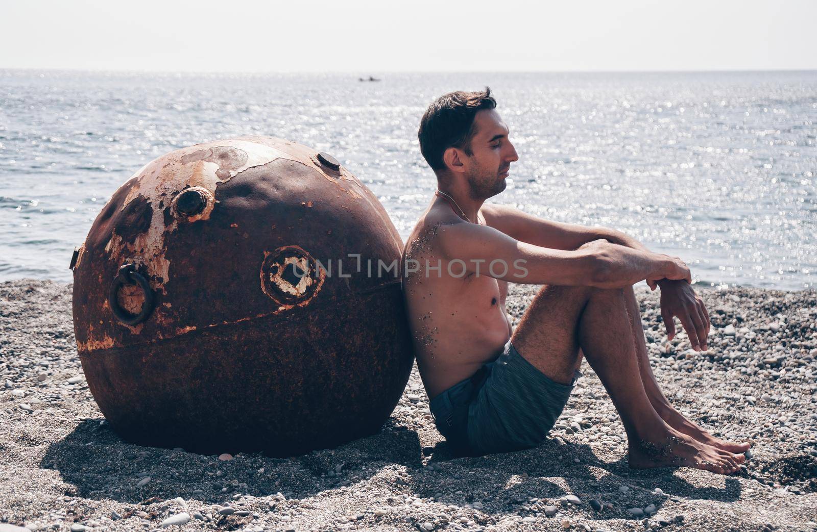 Man doing yoga and meditation outdoors near to old rusty floating marine mine on the beach with rocky shore and sea background. Healthy lifestyle, pollution, nature protection, war and peace concept. by panophotograph