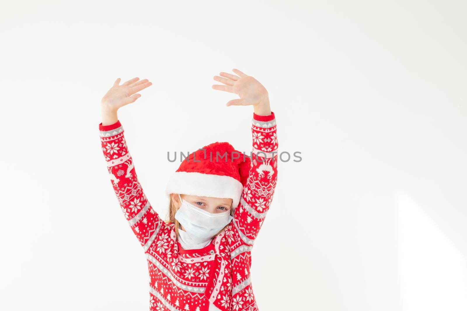 Portrait of a beautiful little girl wearing Santa hat,medical mask and showing hands up, dancing, isolated on white background,copy space. celebrating happy New Year.Child safety social distance by YuliaYaspe1979