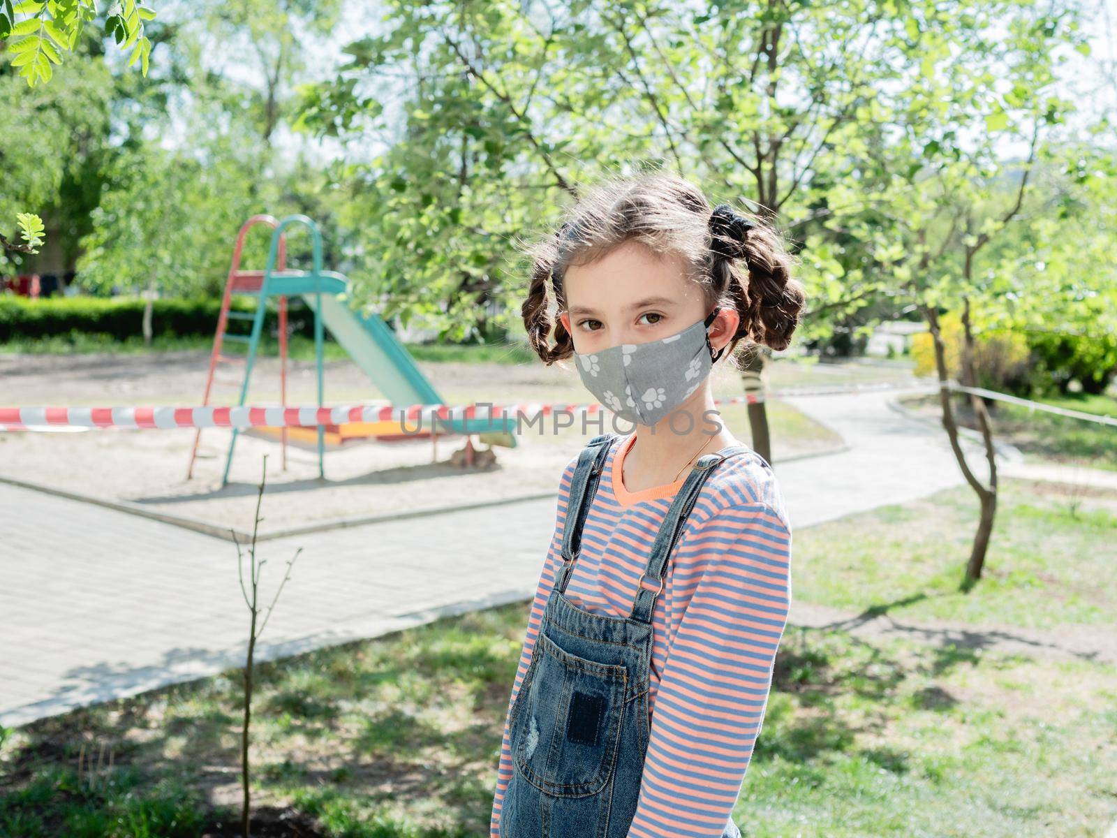 A little girl in a medical mask is not allowed to play in the playground during the coronavirus pandemic. by Utlanov