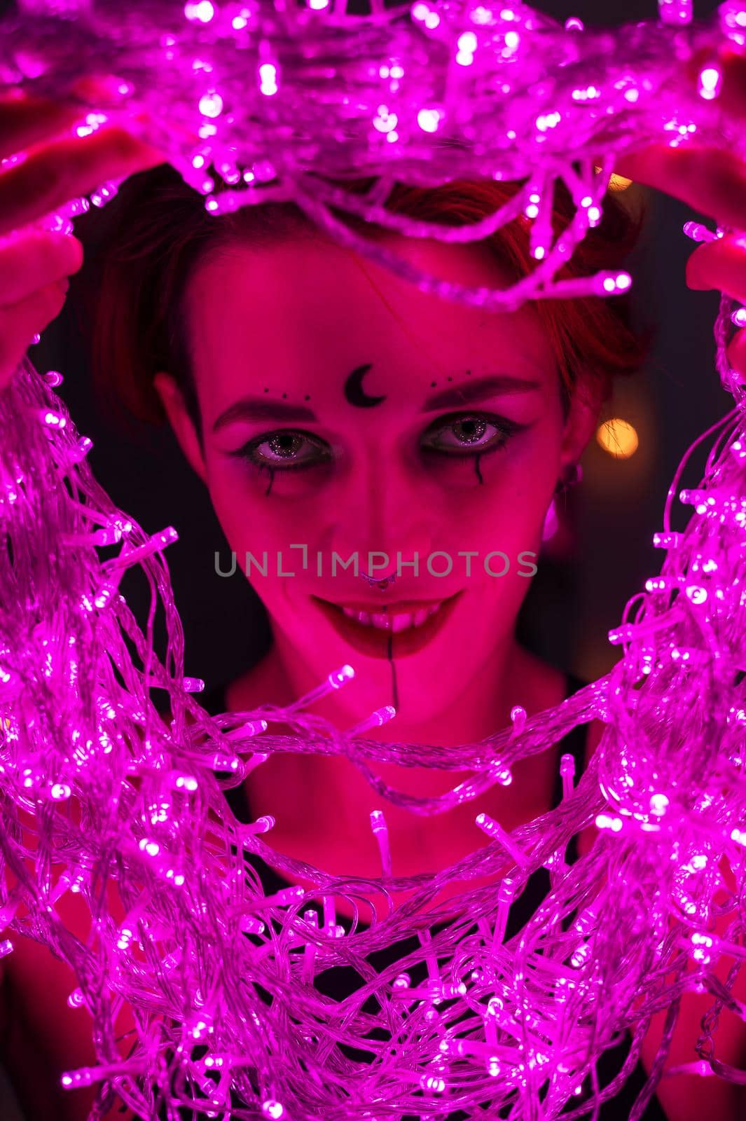 Woman with witch makeup and an earring in her nose. The girl holds a garland of flickering pink lights near her face. Light bulbs illuminate the witch's face in the dark