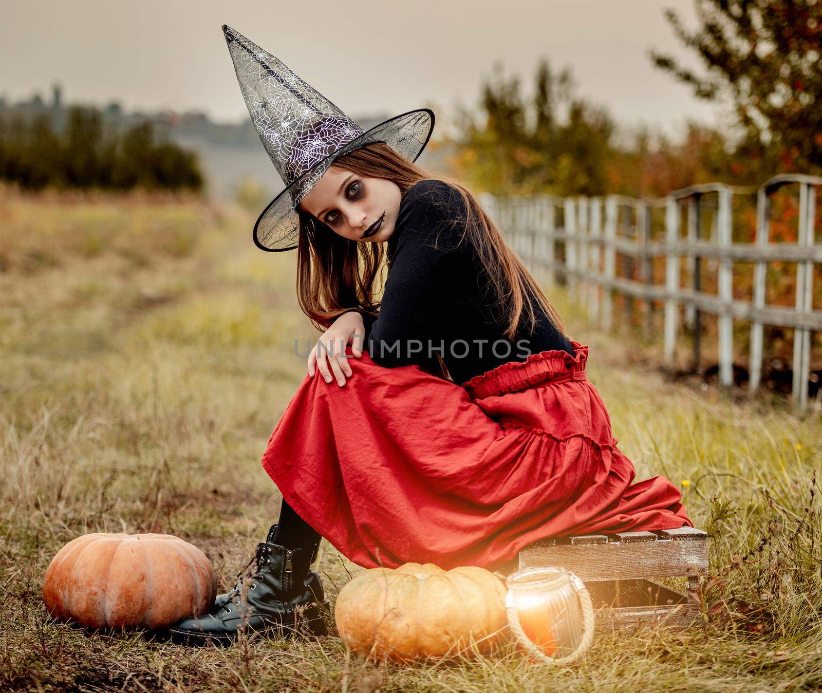 Young girl in halloween makeup and costume sitting on box near pumpkins and candle outdoors