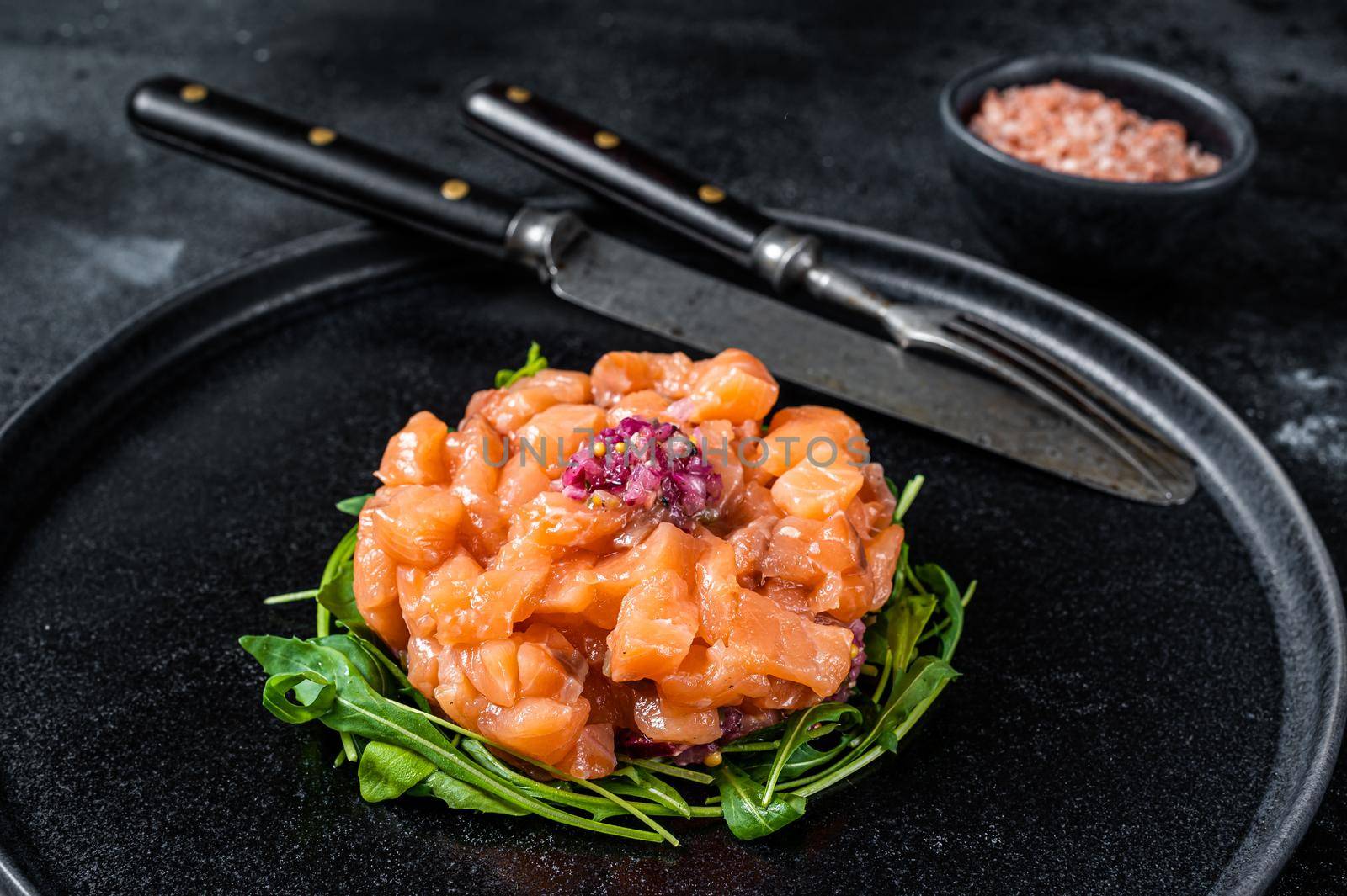 Raw Salmon tartare or tartar with red onion, arugula and capers in black plate. Black background. Top View.