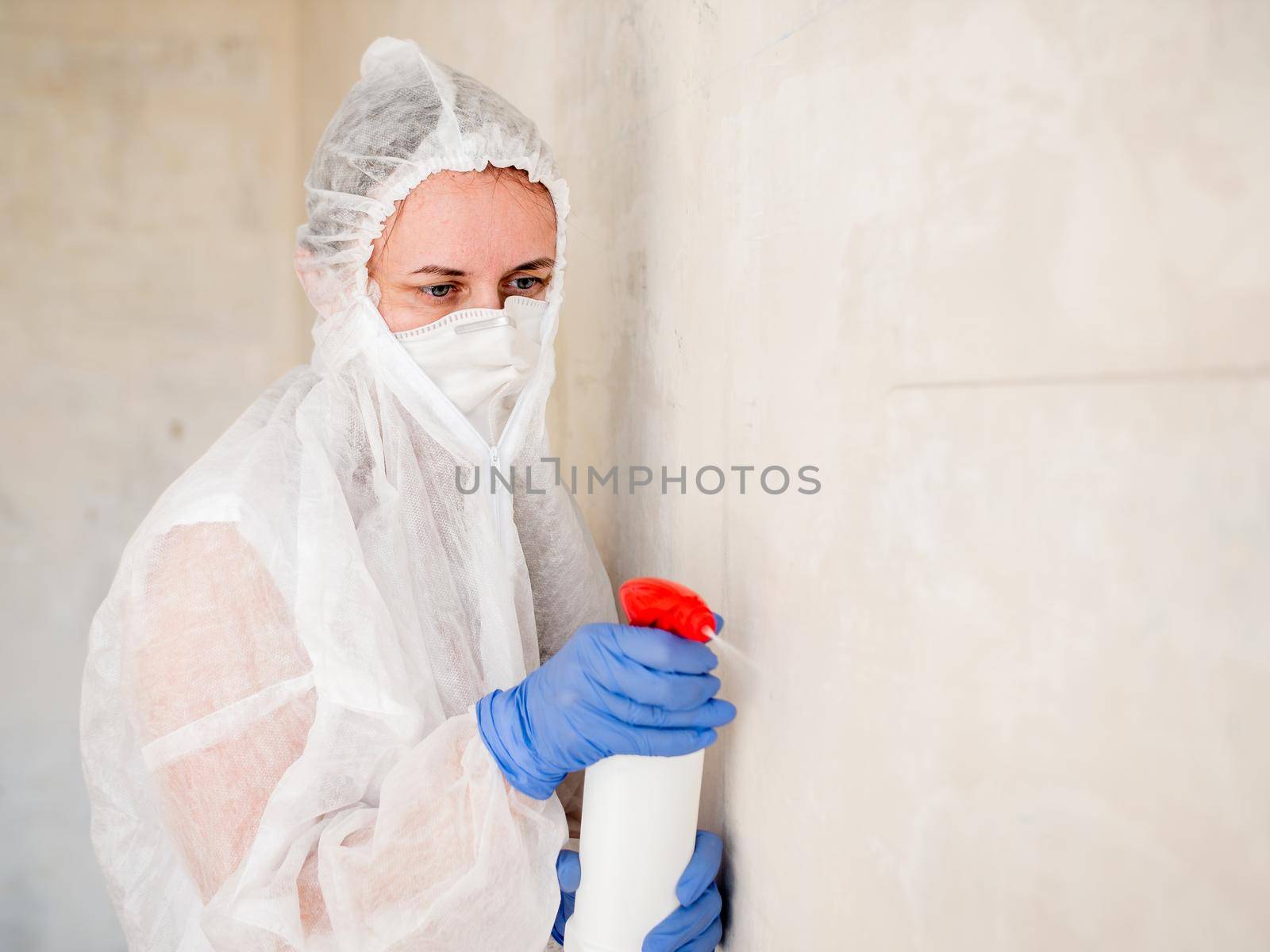A woman in a protective suit sprays the walls of an apartment with a chemical agent to remove mold.