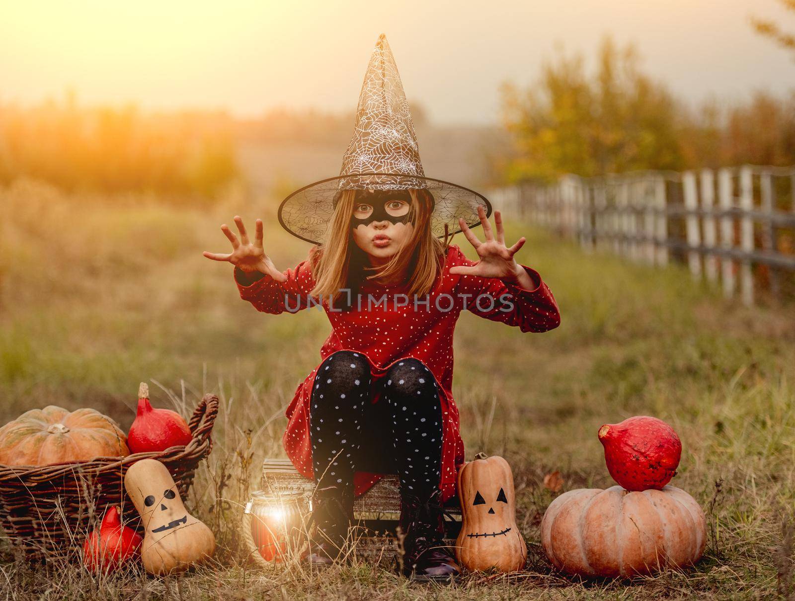 Little girl in halloween costume sitting next to pumpkins while frightening at camera on autumn nature