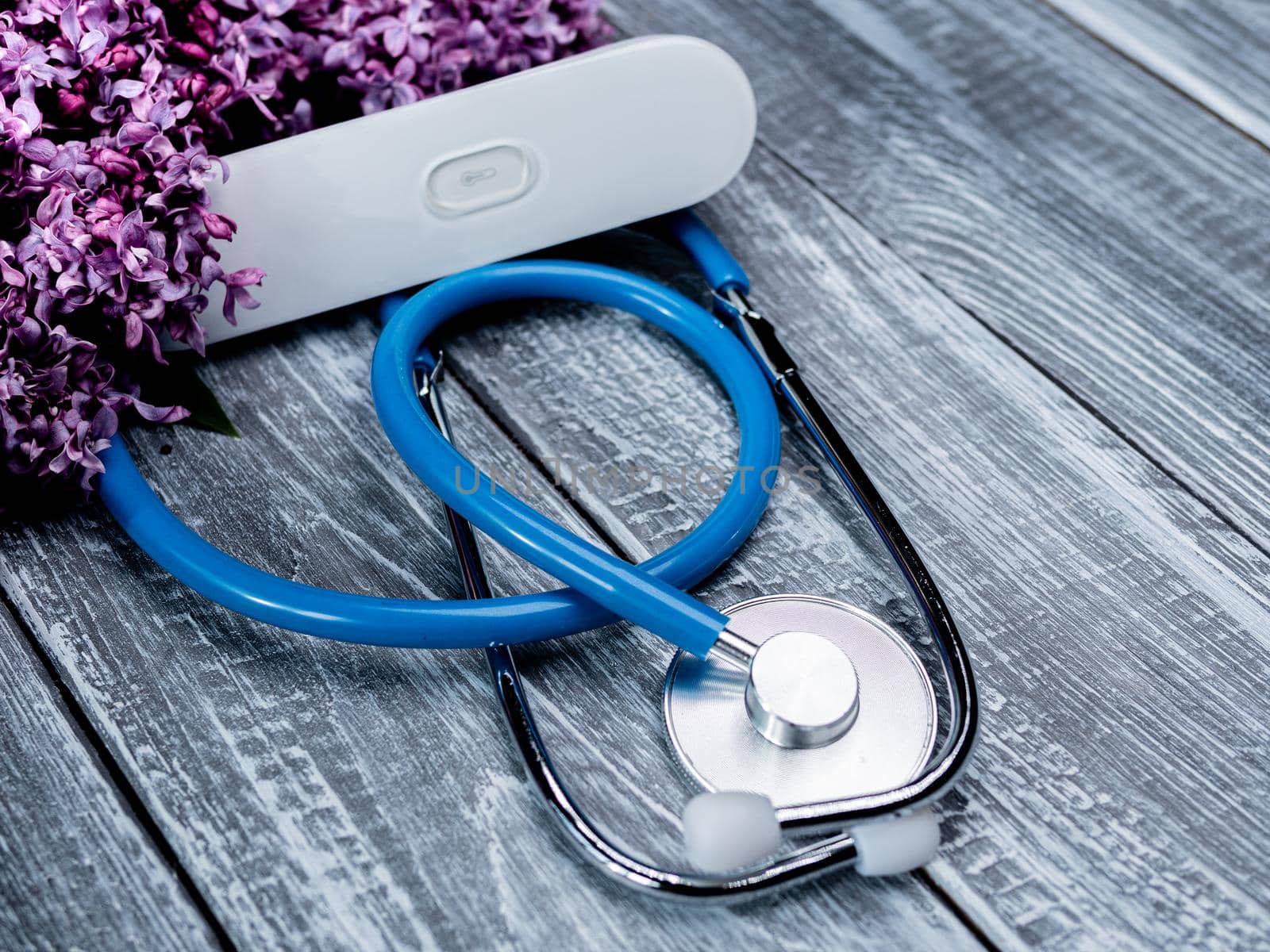 A stethoscope and an electronic thermometer lie on a wooden background next to a bouquet of lilacs. Concept spring and the coronavirus pandemic