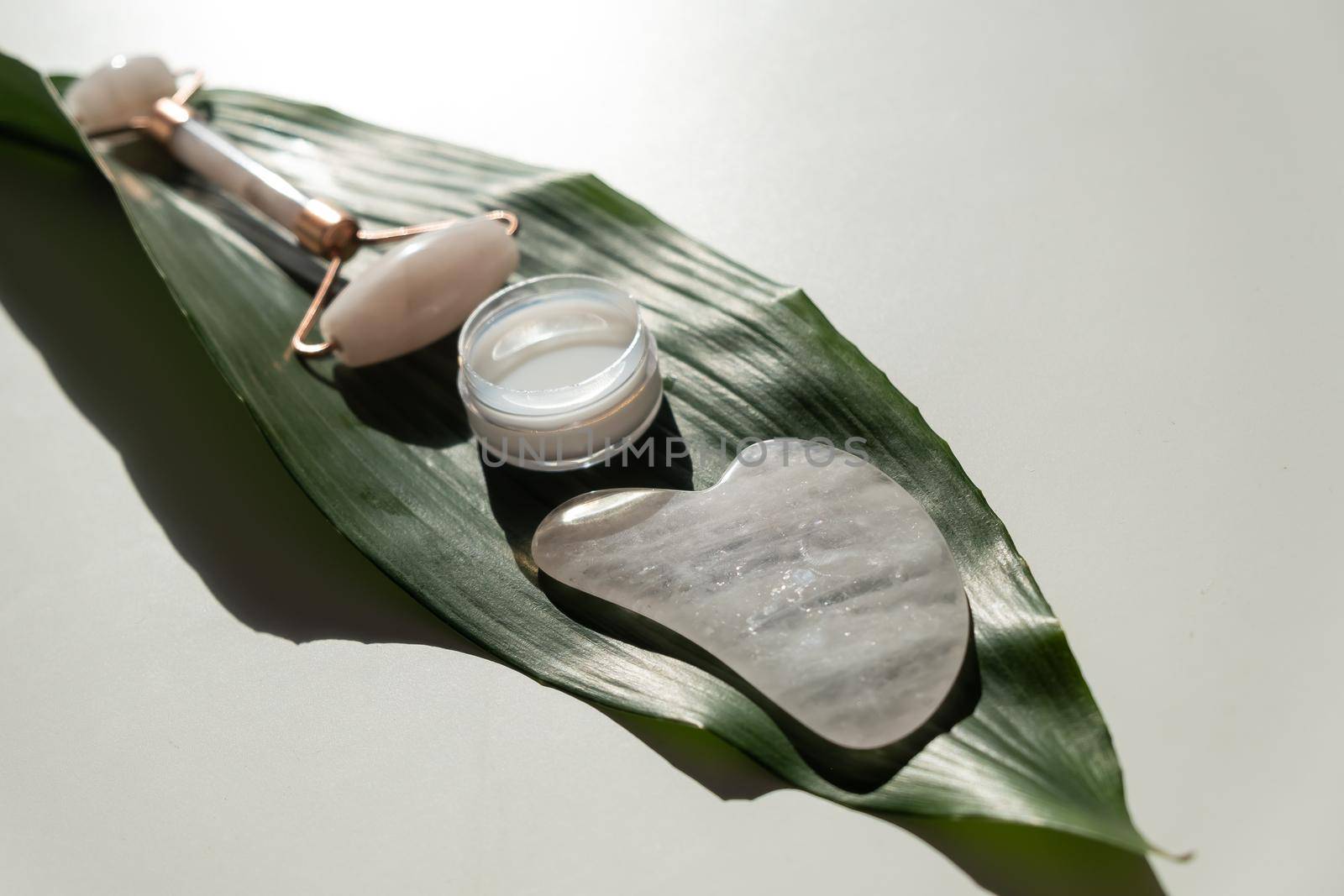 Pink gouache scraper, roller, jar of green leaf cream on a white table. Equipment for self-massage and skin care for the face and neck.