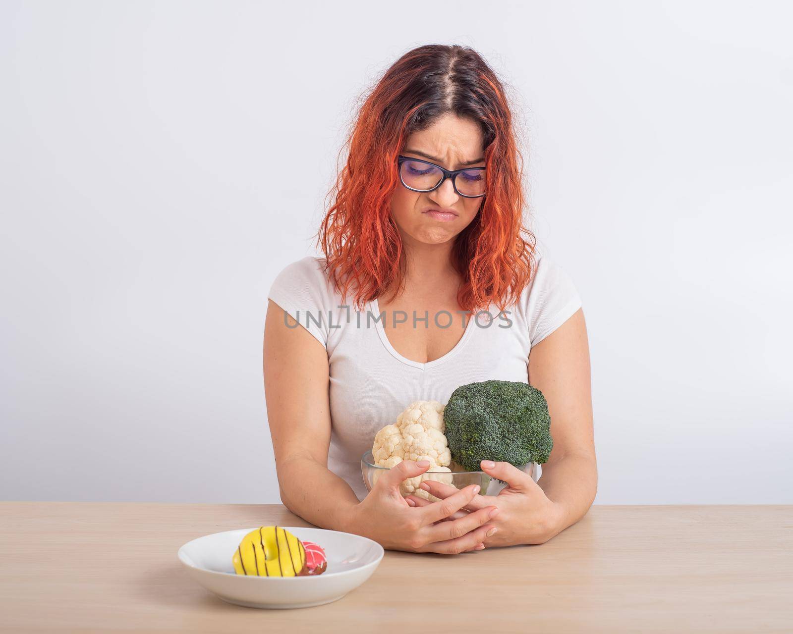 Caucasian woman on a diet dreaming of fast food. Redhead girl chooses between broccoli and donuts on white background. by mrwed54