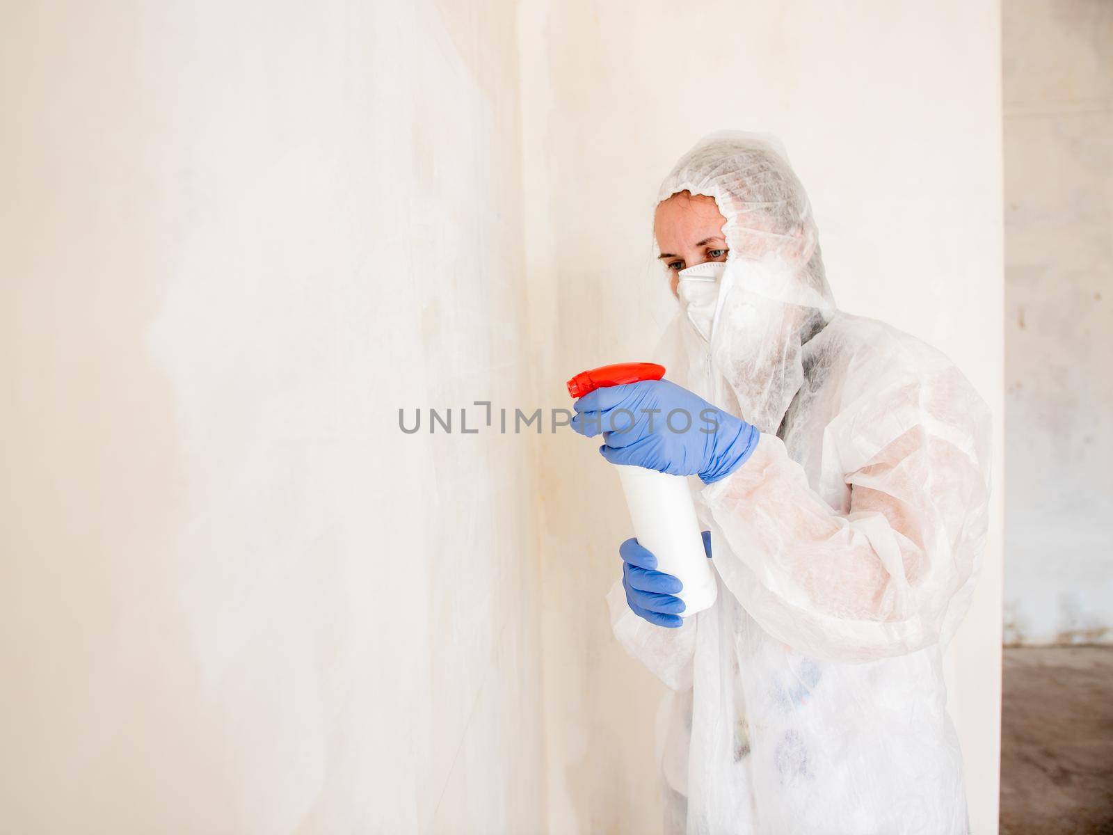 A woman in a protective suit sprays the walls of an apartment with a chemical agent to remove mold. by Utlanov