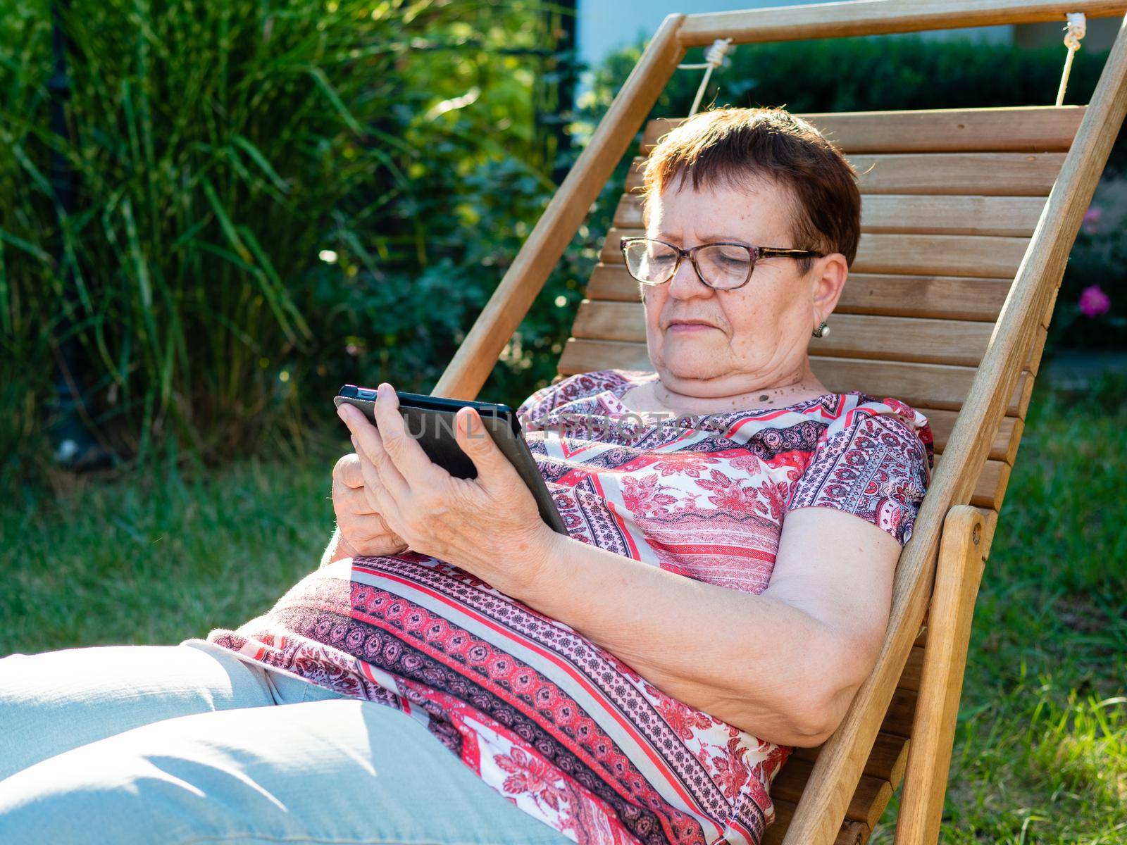 An elderly woman with glasses reads an e-book sitting on a wooden chaise longue in the backyard of a house. by Utlanov