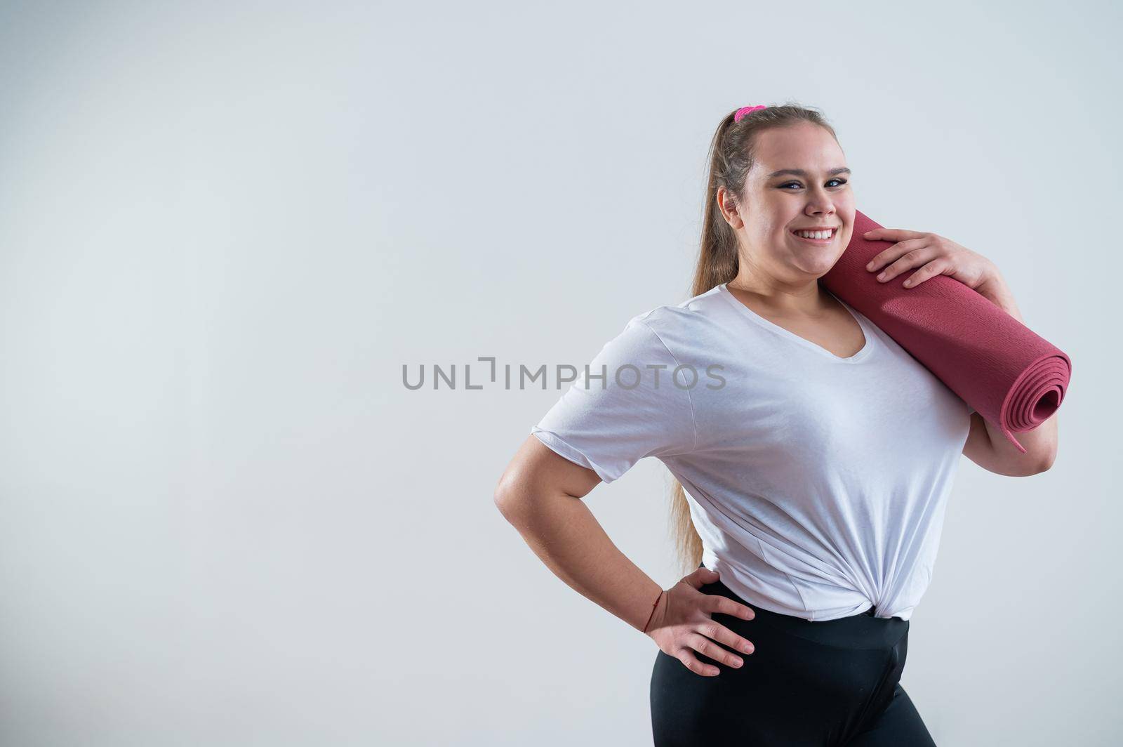 Young fat caucasian woman holding a sport mat. Charming plus size model in sportswear stands on a white background.