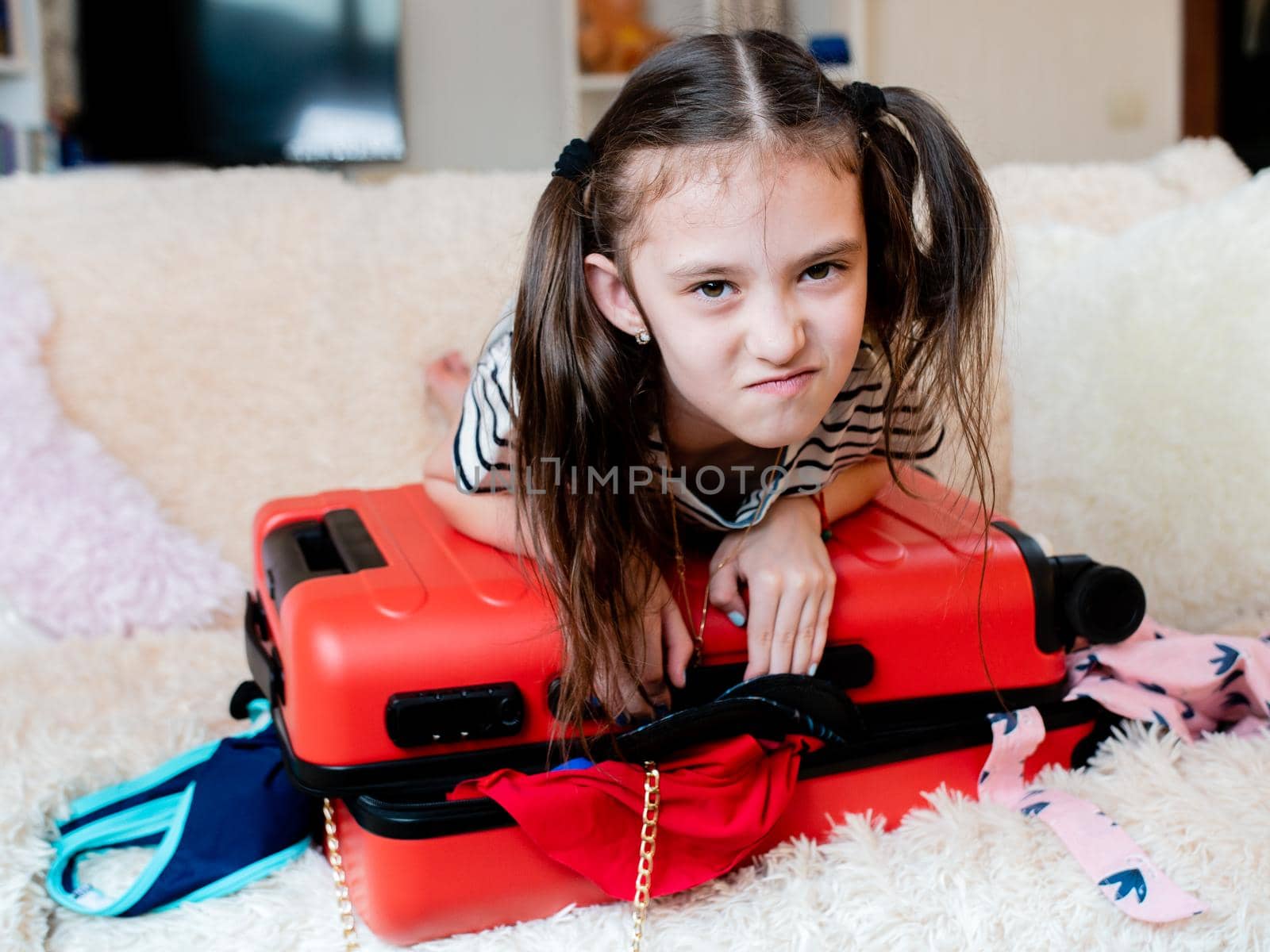 little girl leaned on top of the suitcase trying to close it. A suitcase full of things does not close. by Utlanov