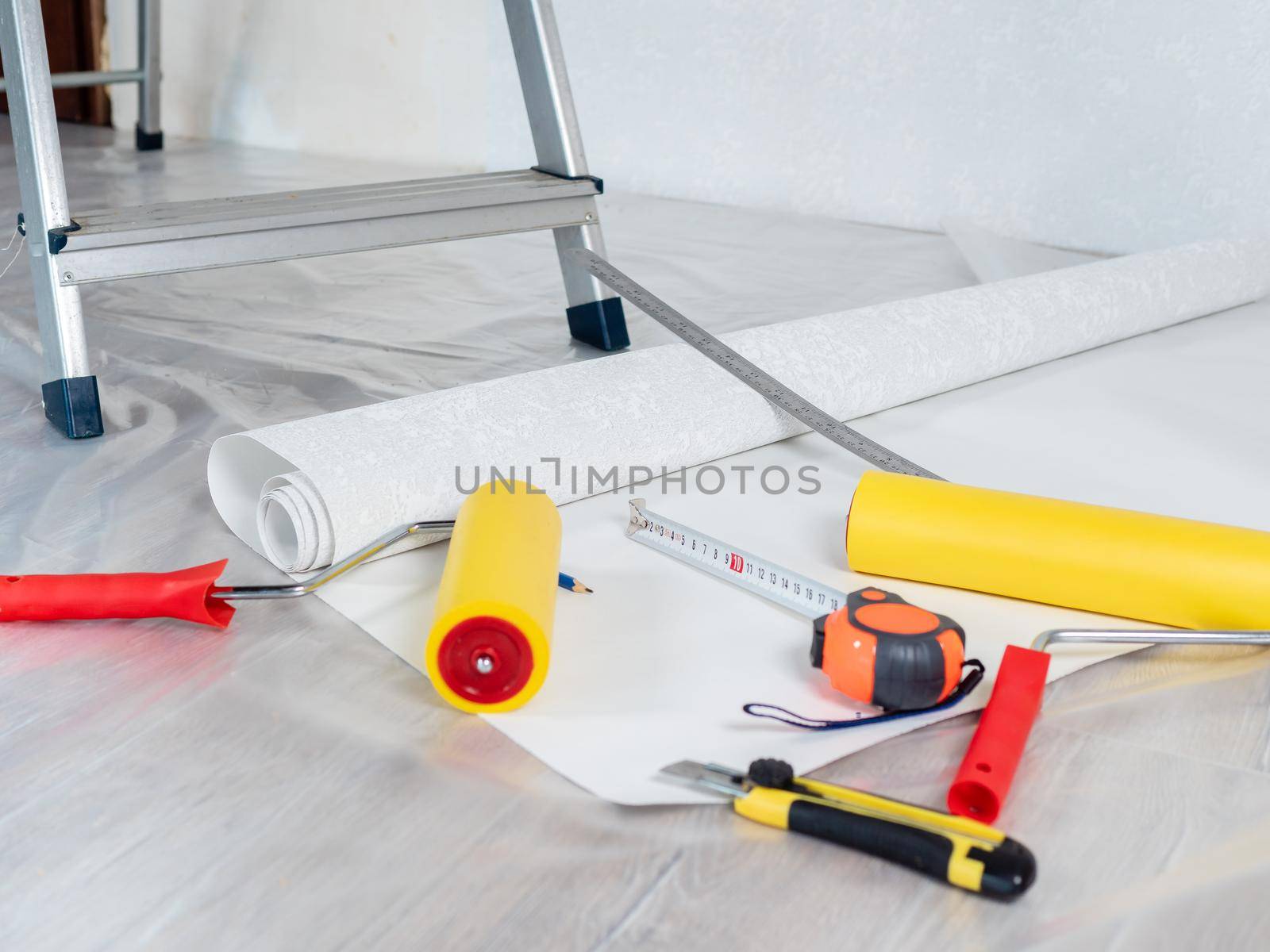 A knife and tape measure lie on the floor next to a stepladder and rolls of Wallpaper for repairs in the apartment, close-up. by Utlanov