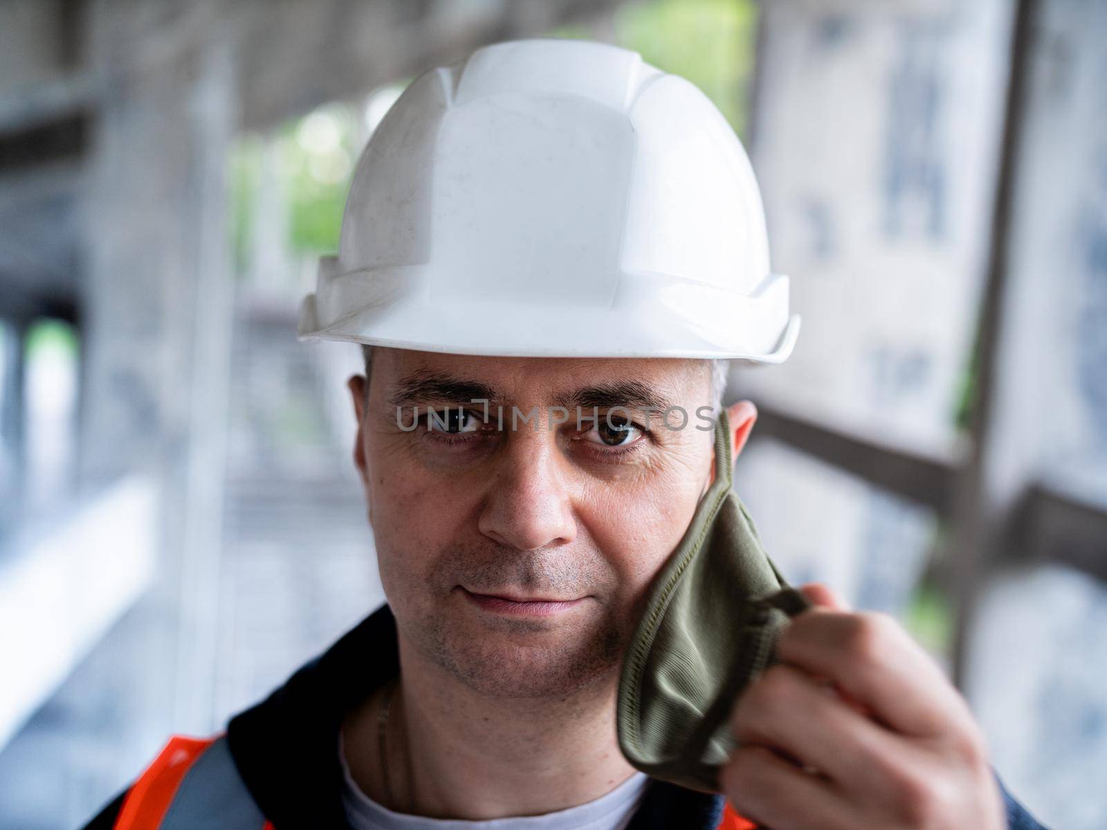 The man removes the protective mask from his face to symbolize the end of the pandemic. Portrait of a male Builder in a construction helmet and a medical antiviral mask.