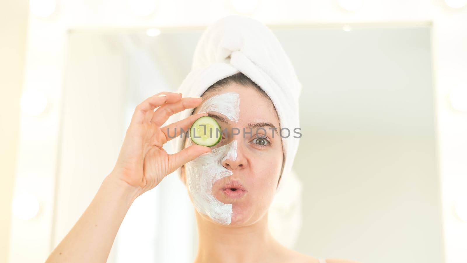 Cheerful woman with a towel on her hair and in a clay face mask fooling around with cucumbers in her hands. Taking care of beauty at home by mrwed54