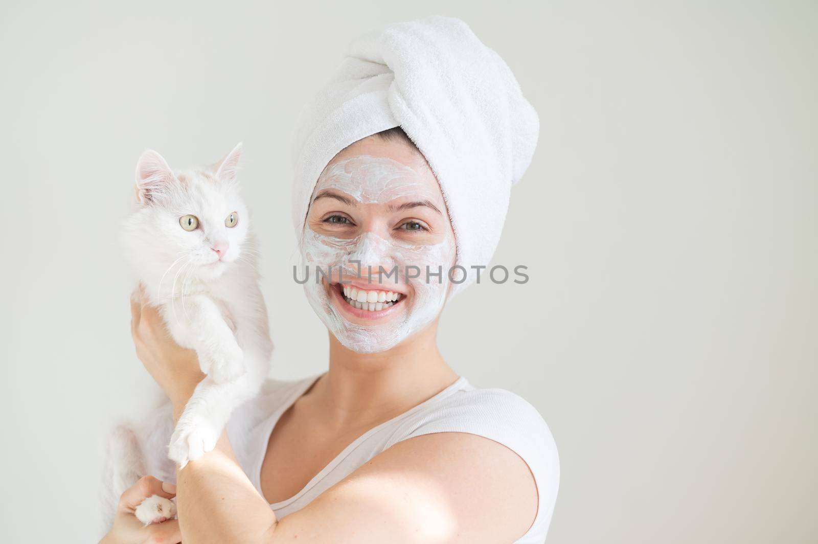 A woman with a towel on her hair and a clay mask on her face is holding a white fluffy cat on a white background. Copy space.