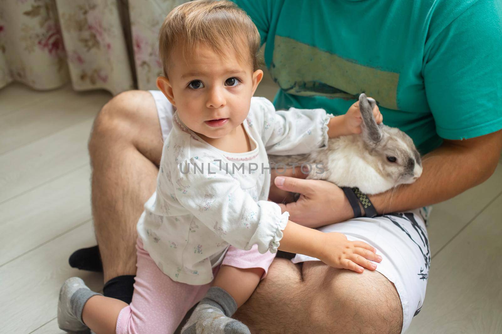 adorable baby sits in dad's arms and strokes a decorative rabbit. father shows little child easter bunny. domestic animals in a family with children