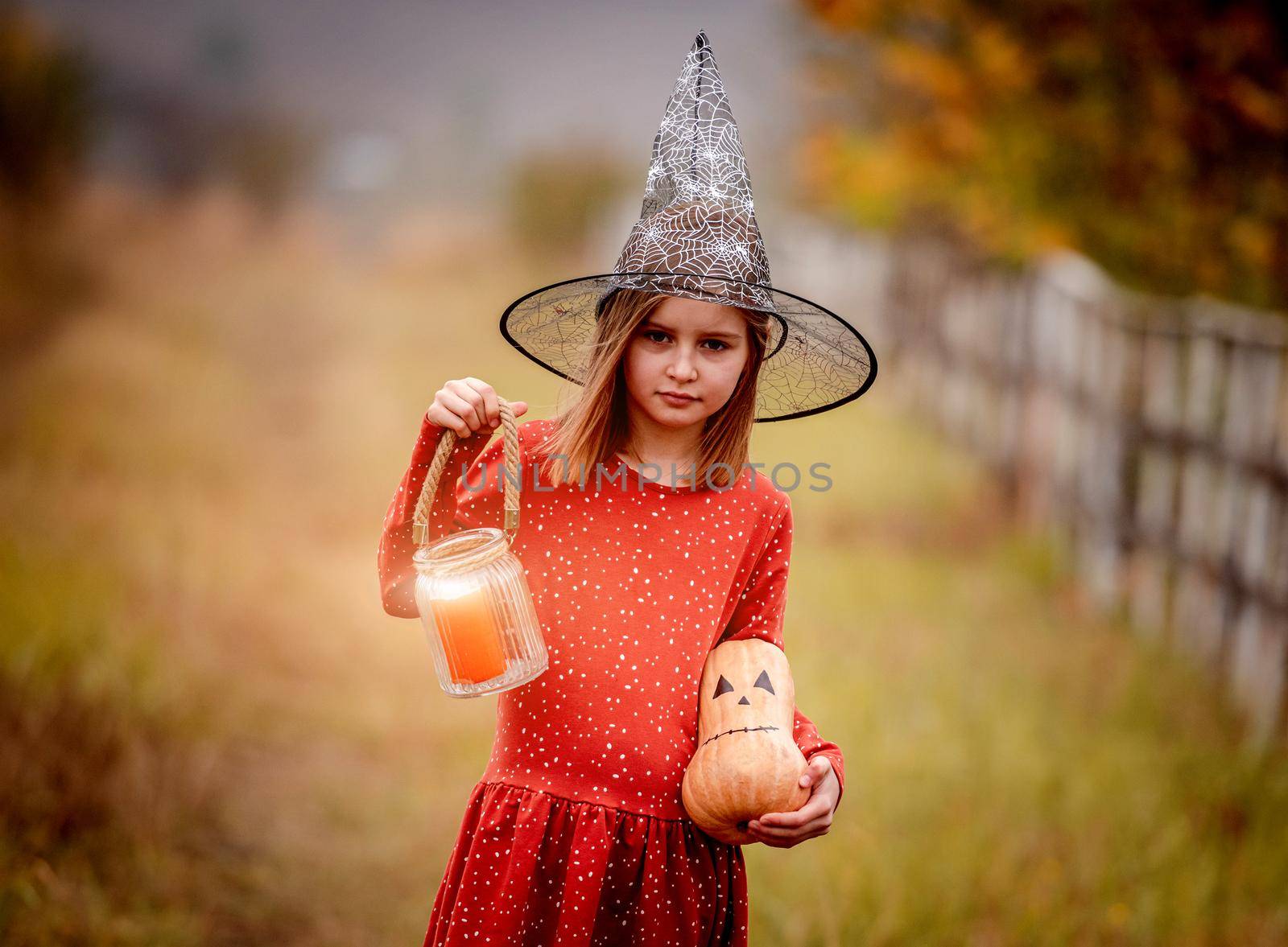 Little girl in witch hat holding candle and pumpkin standing on nature near flying bats