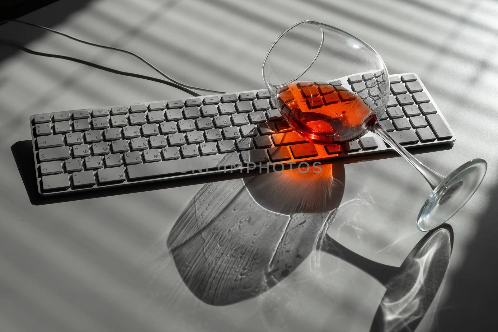 A glass of red wine lies on the keyboard on a white table with a shade from the blinds.