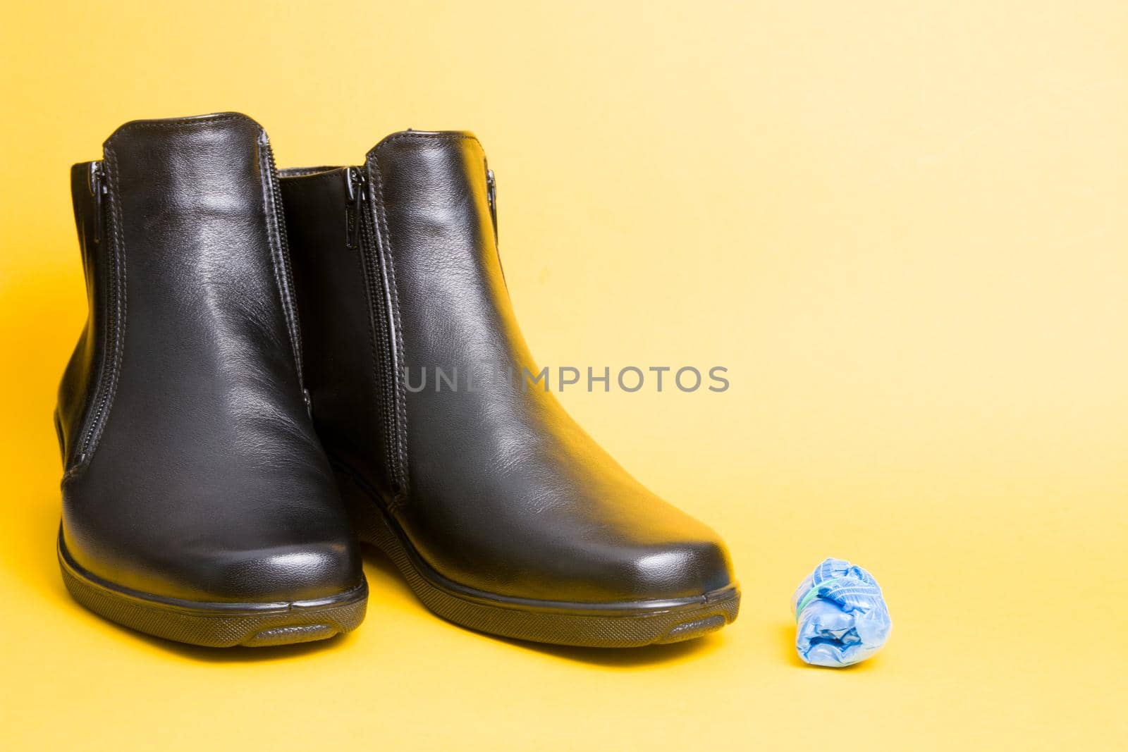 black leather boots and blue disposable shoe covers on a yellow background copy space