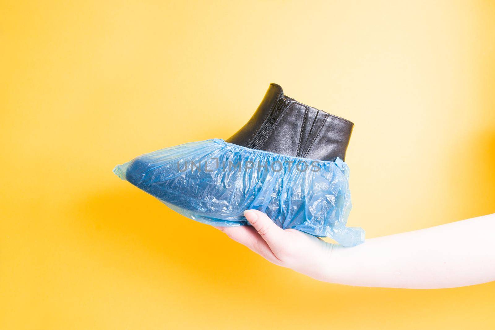 female hand holds a black leather female boot in boot covers, yellow background