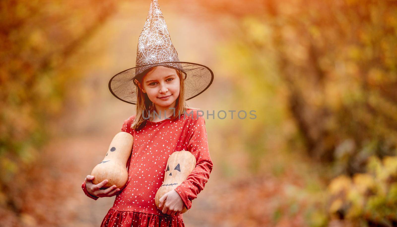 Little girl in witch hat holding decorated pumpkins for halloween on colorful autumn nature background