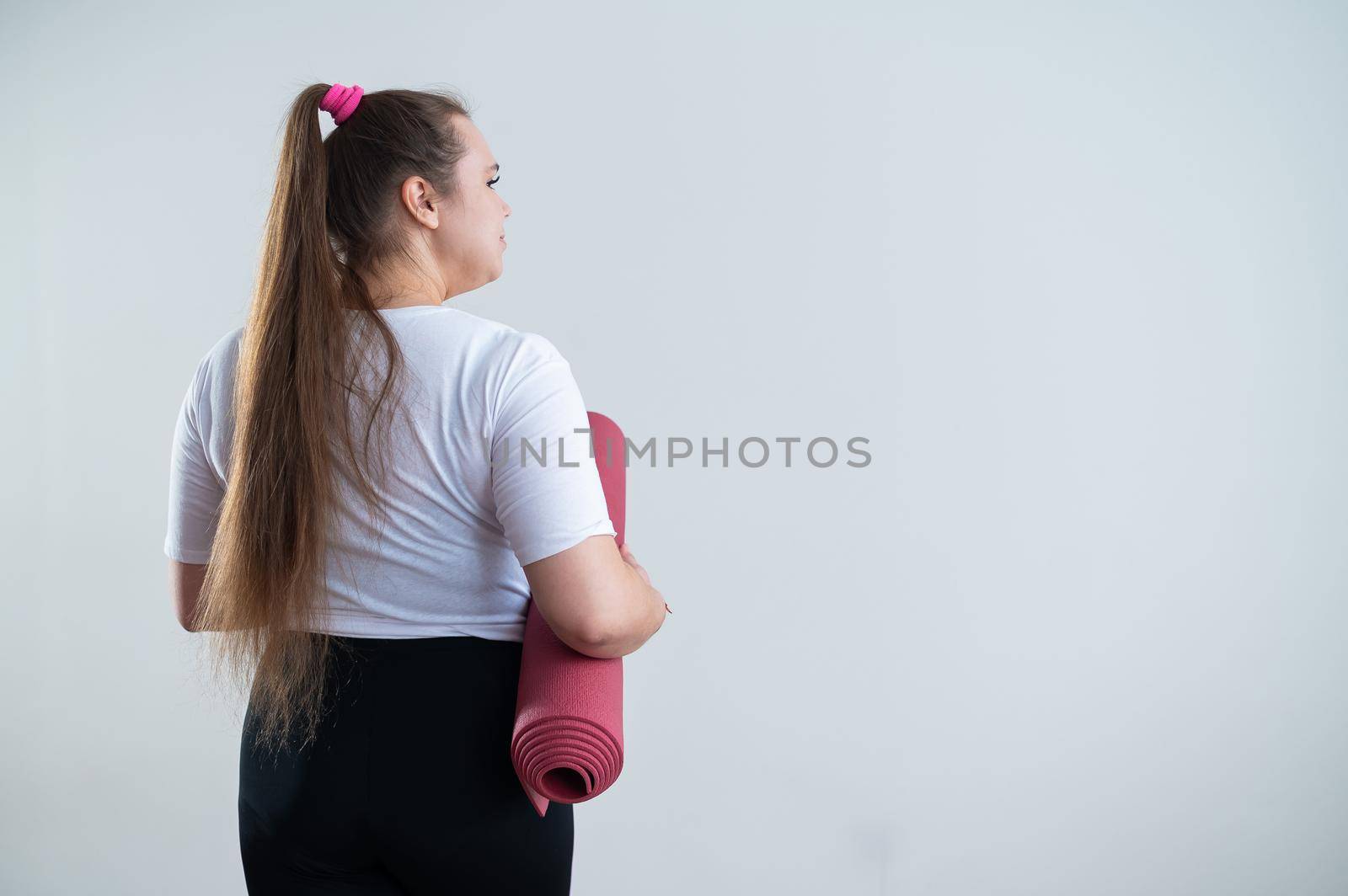 Young fat caucasian woman holding a sport mat. Charming plus size model in sportswear stands on a white background.