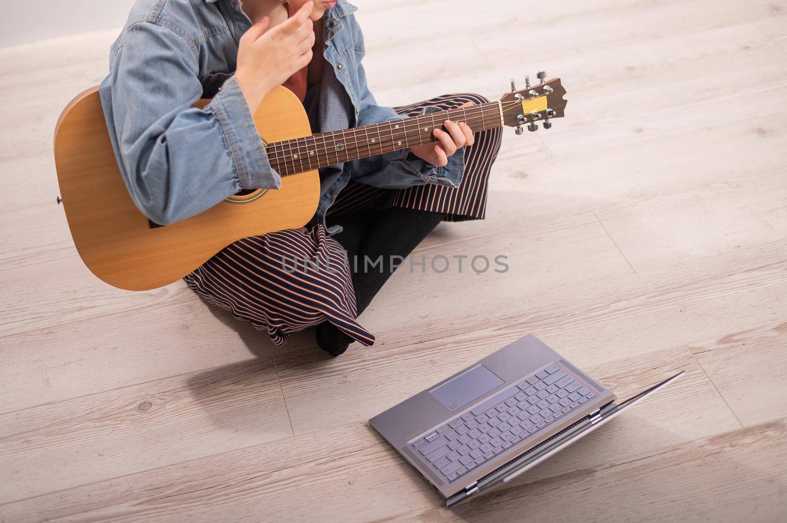 Young hipster woman sitting on the floor at home and watching guitar tutorials on laptop. Distance learning music in quarantine by mrwed54