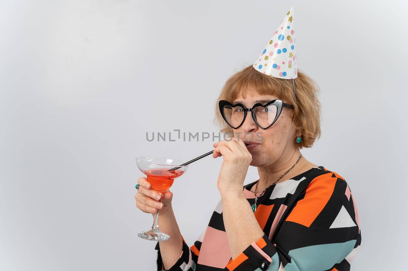 Portrait of a smiling elderly woman in a festive cap and glasses with hearts drinking a cocktail on a white background.