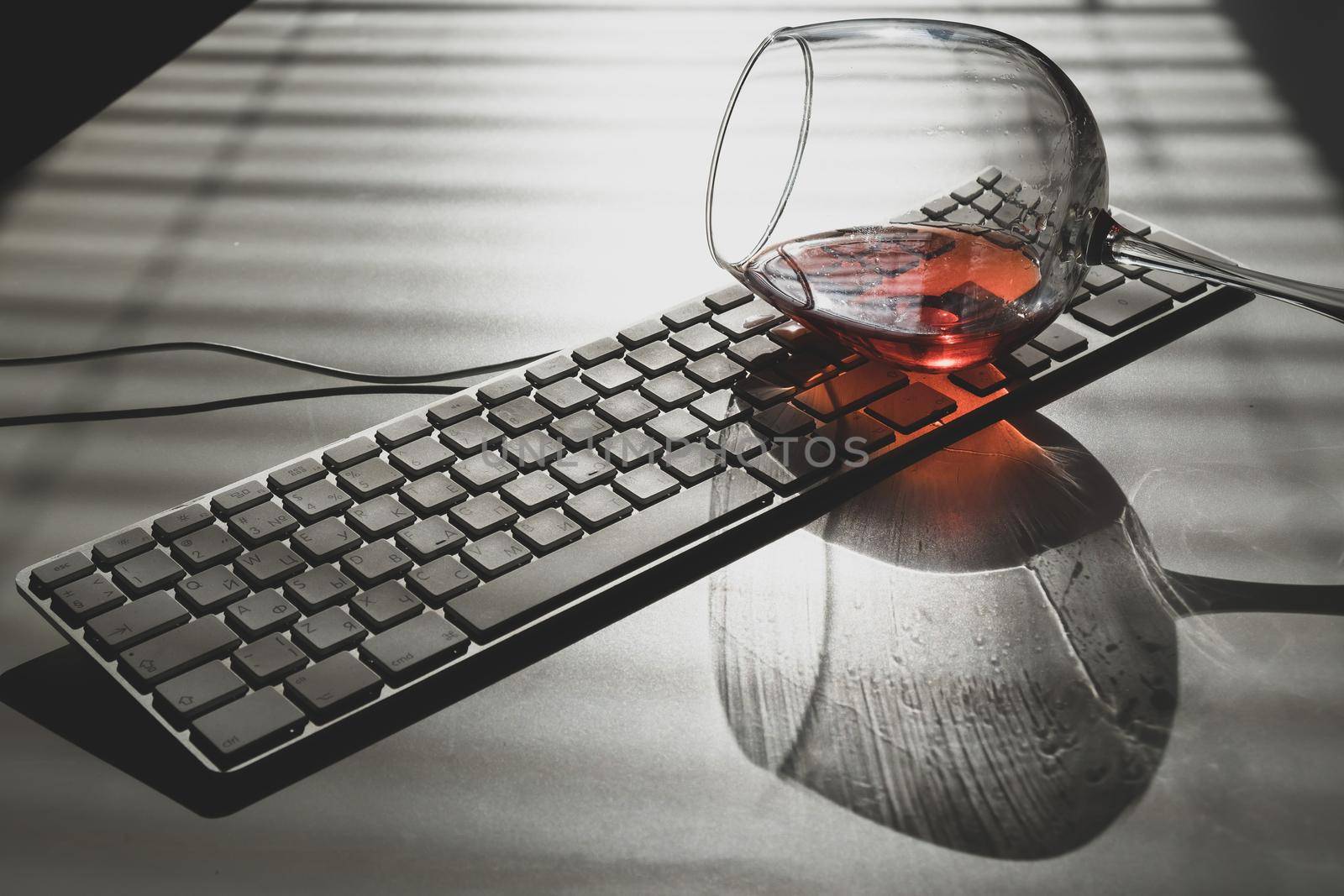 A glass of red wine lies on the keyboard on a white table with a shade from the blinds by mrwed54