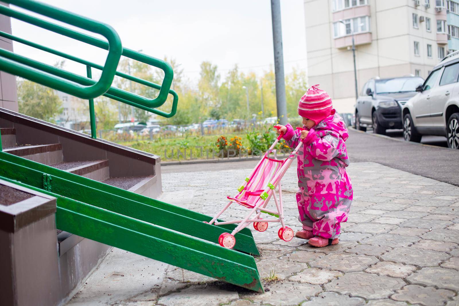 cute toddler with a toy stroller walks along steel railing ramp for wheelchair, carts and strollers. gentle descent from the stairs outdoors