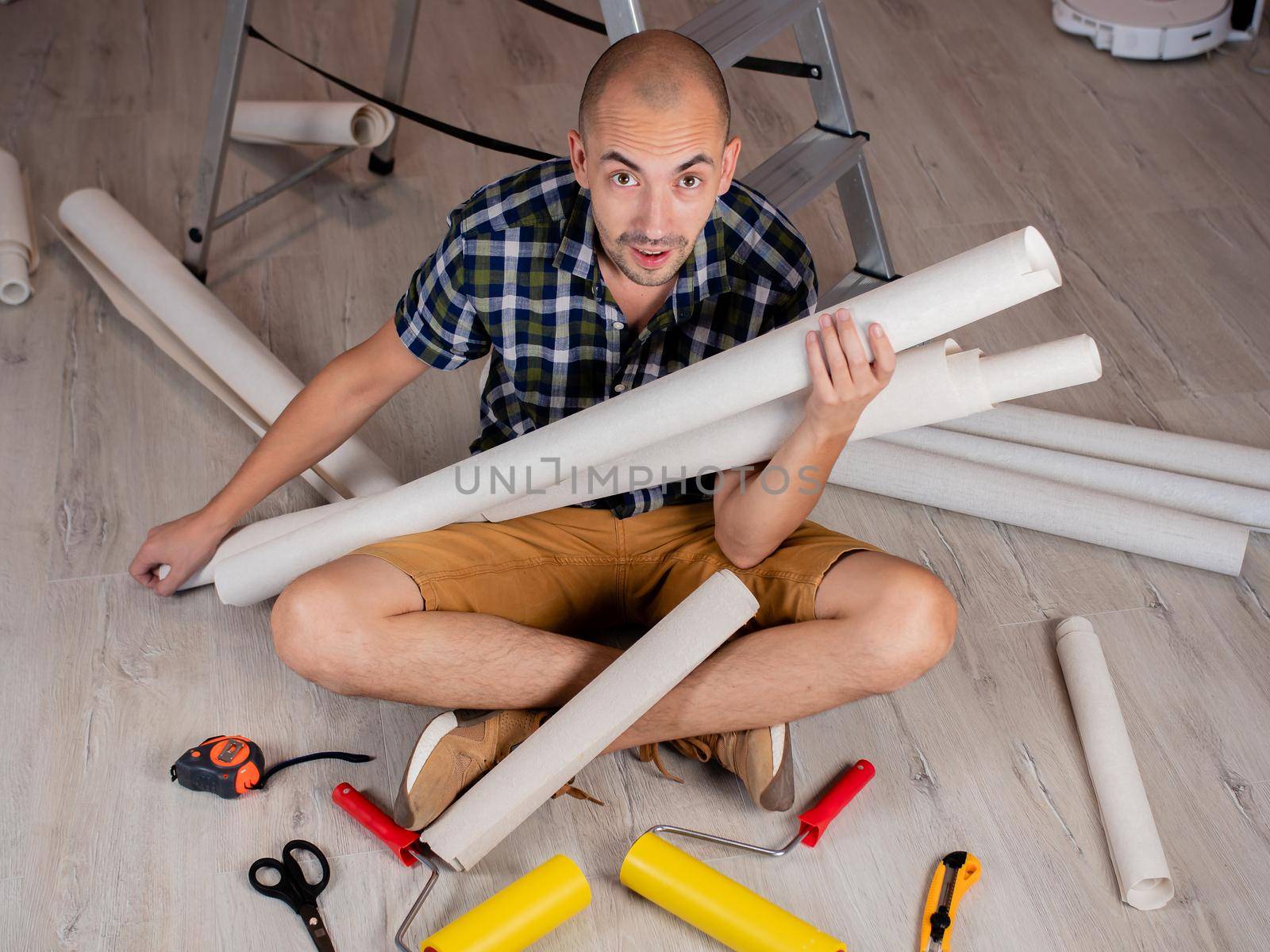 A man is sitting on the floor with many rolls of Wallpaper and tools for gluing them. by Utlanov