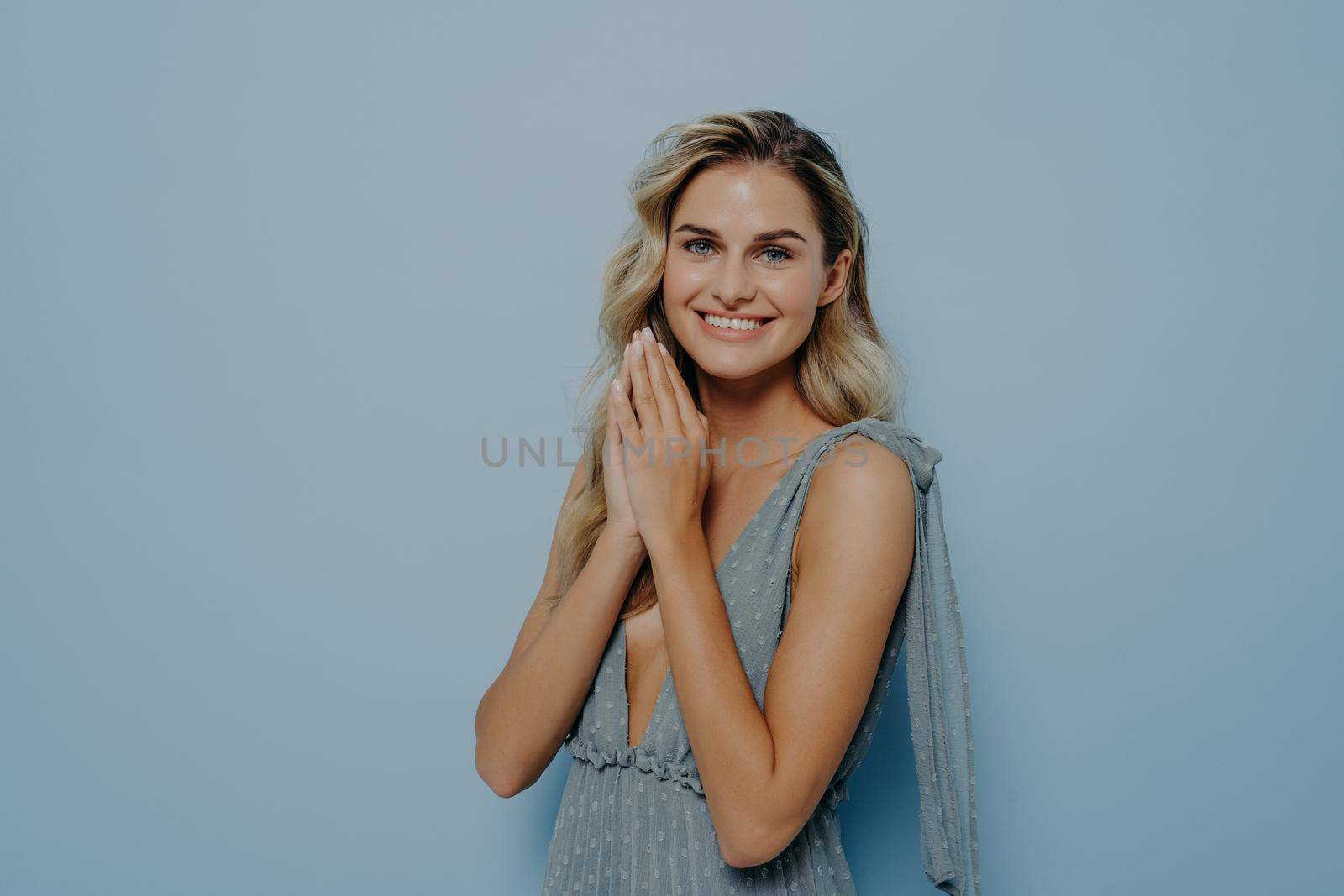 Friendly beautiful young blonde woman wearing romantic summer dress with trustworthy angel stance, holding hands in praying gesture, smiling and asking for help, standing alone next to blue background