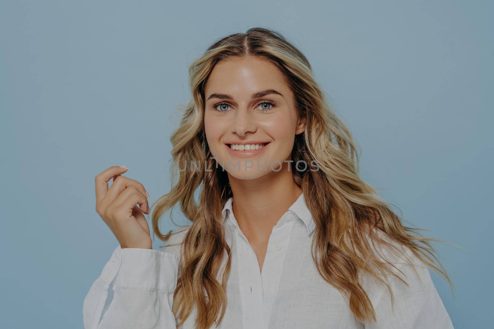 Headshot of beautiful caucasian female smiling broadly at camera demonstrating her white teeth, happy woman with perfectly styled hair and radiant healthy skin isolated against blue studio background