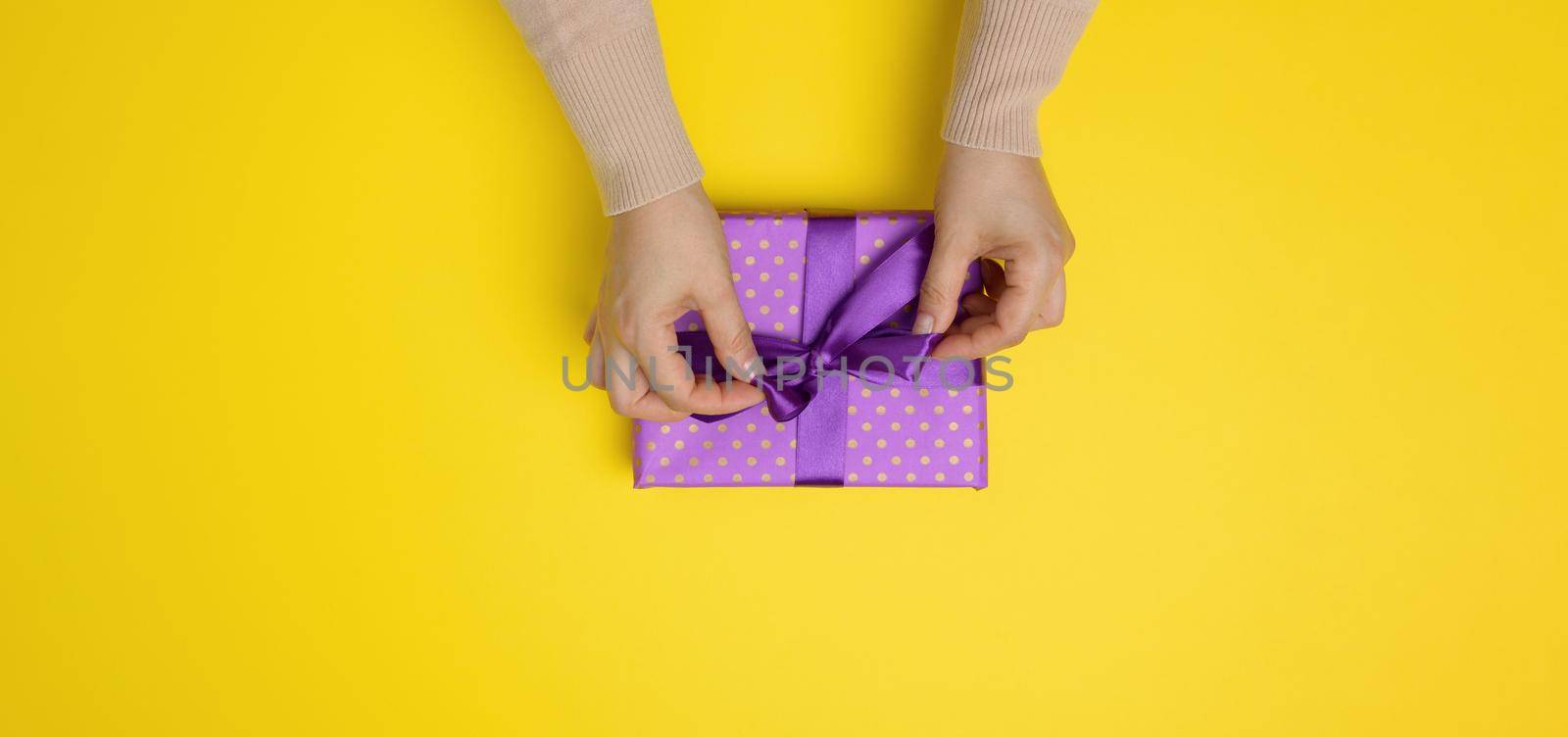 female hands tie a silk ribbon on a wrapped box in purple paper. Yellow background, top view