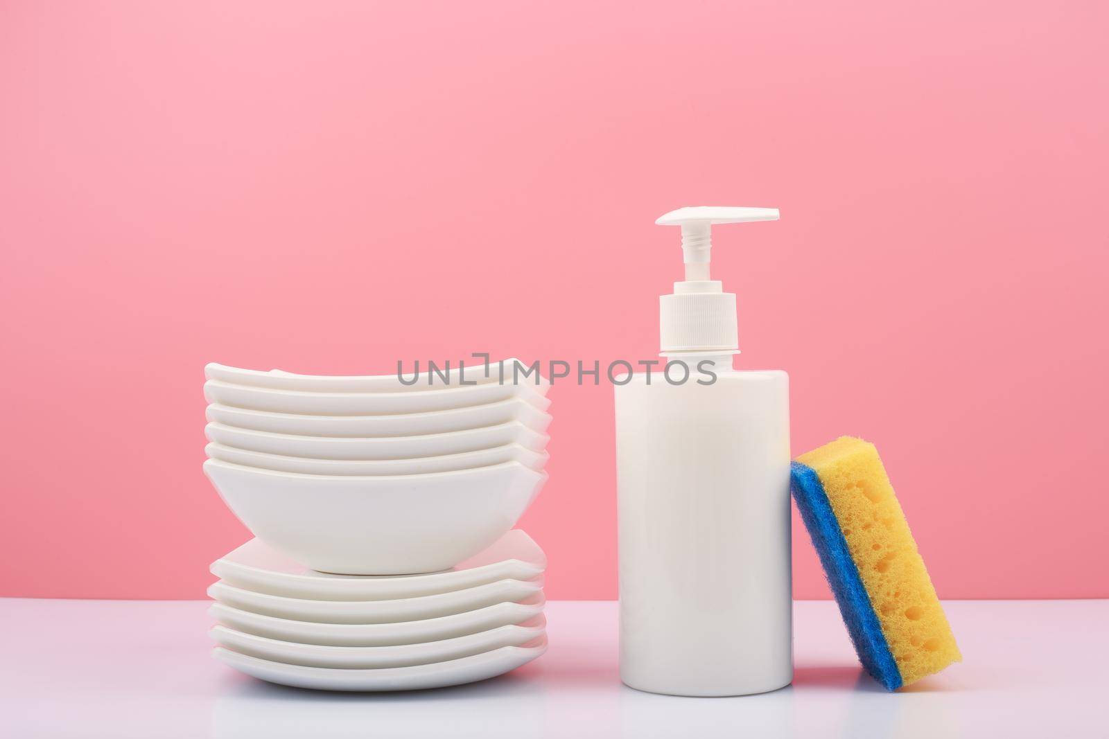 Dishwashing and housework concept. Clean white ceramic plates, detergent in white plastic tube andyellow cleaning sponge against bright pink background. 