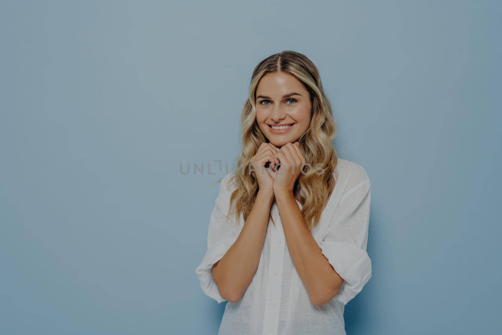 Cute young blonde woman with wavy perfect hairstyle folded hands under her chin and waiting for desired gift in excitement, dressed in white shirt. Happy facial expression and positive emotions