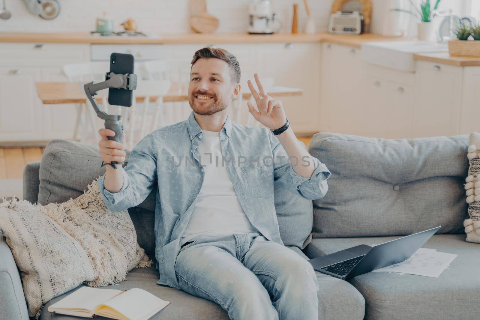 Positive young man showing peace sign during video call while sitting on couch by vkstock