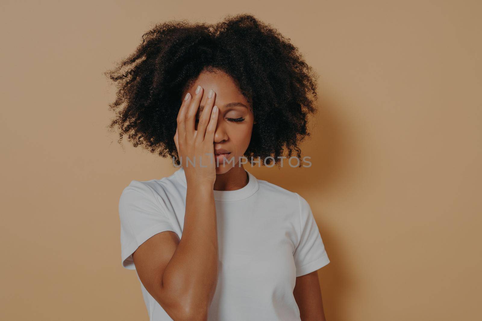 Dark skinned female covering face with hand and feeling anxiety or depression against beige wall by vkstock