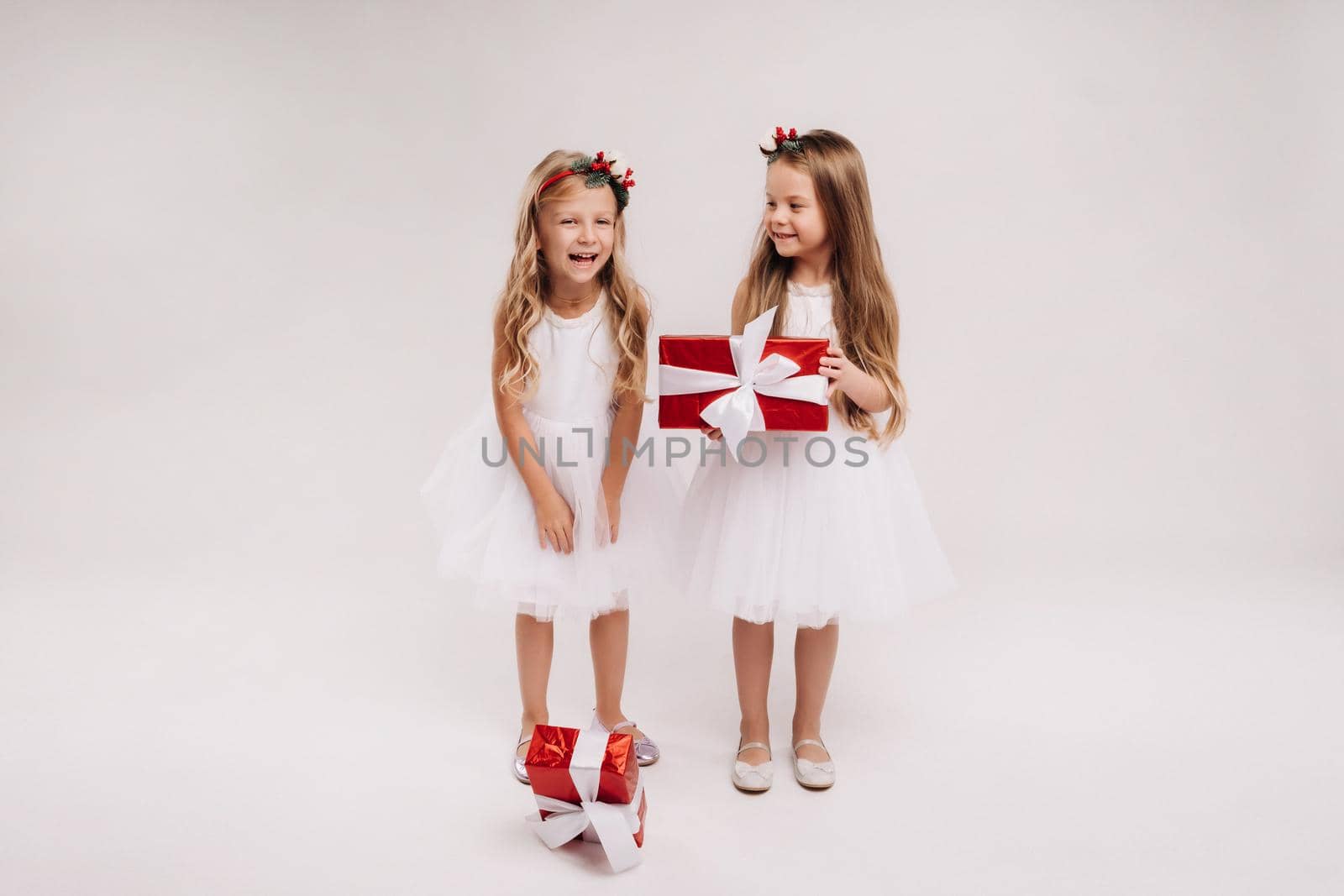 two little girls in white dresses with Christmas gifts on a white background smile.