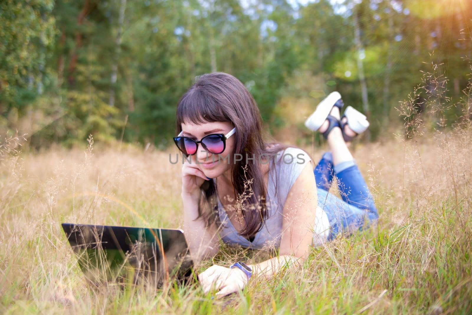 happy young woman with laptop on a sunny lawn. freelancer works in nature. student studies remotely by Mariaprovector