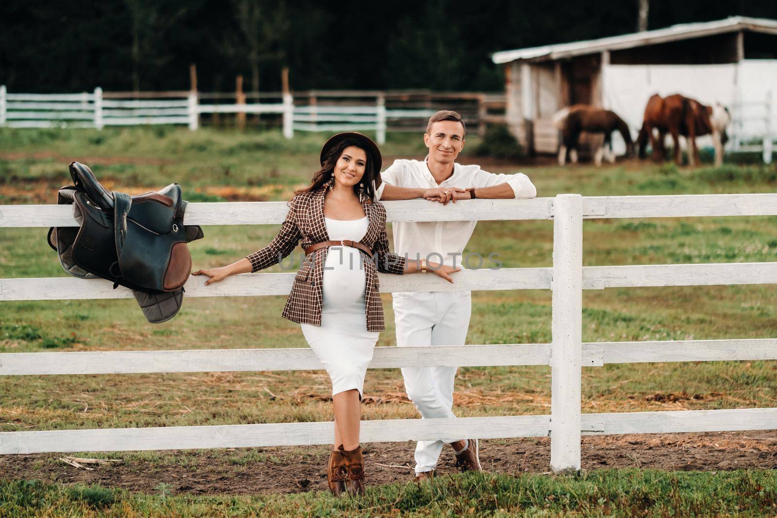 a pregnant girl in a hat and her husband in white clothes stand next to the horse corral.a stylish couple waiting for a child stand on the street near the horse corral