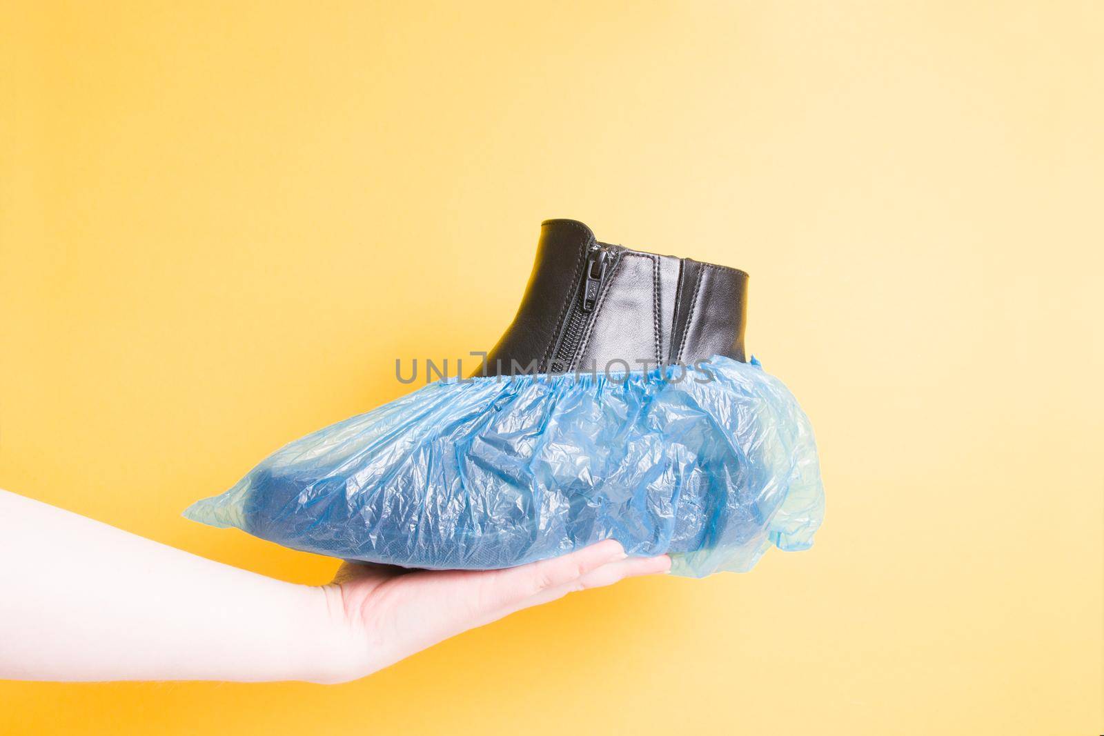 female hand holds a black leather female boot in boot covers, yellow background