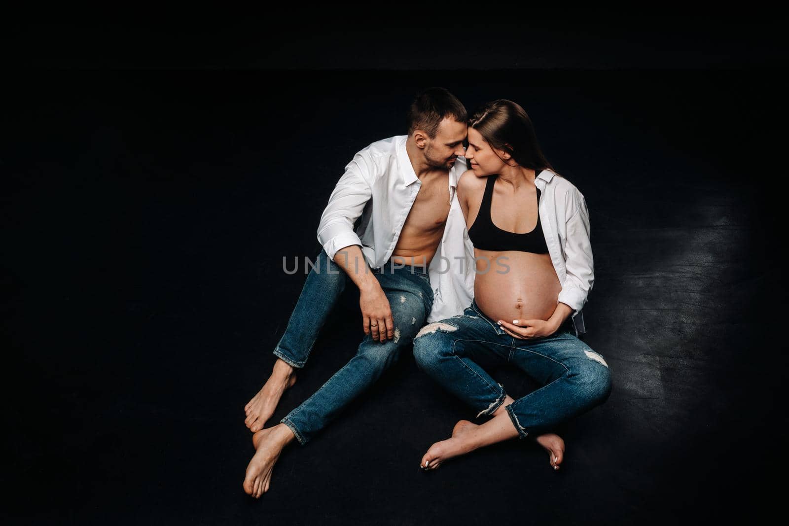 a pregnant woman and a man in a white shirt and jeans in a studio on a black background by Lobachad